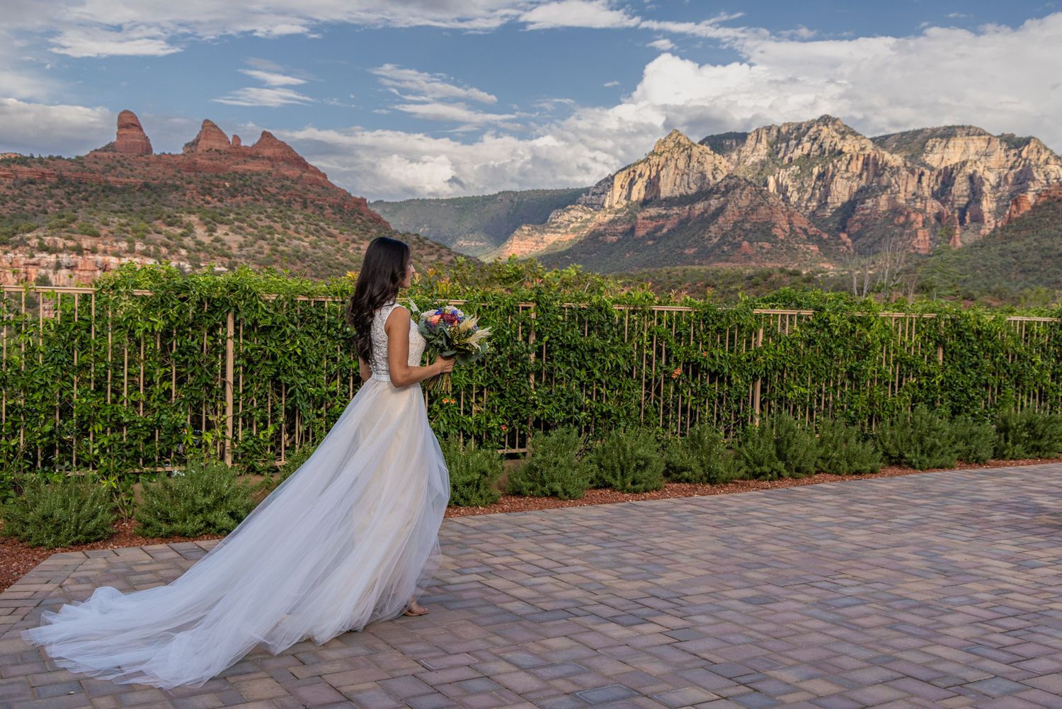 Bride in flowing white gown holding bouquet stands on patio overlooking lush vineyard with red rock Sedona mountains backdrop.