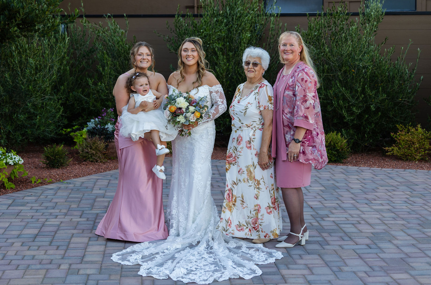 Bride with bouquet poses with bridesmaids and family members in garden setting.