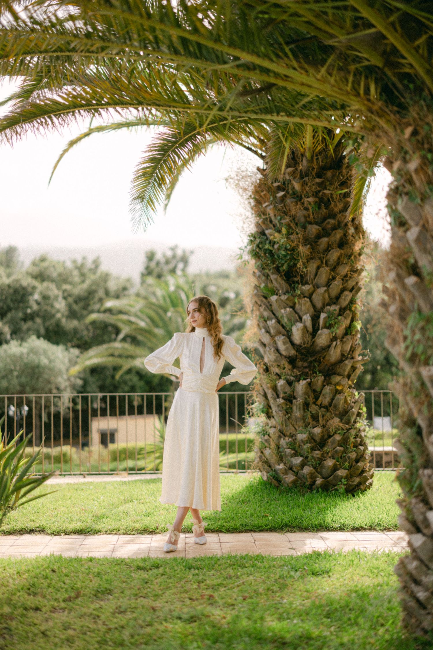 Woman in white midi dress standing among tall palm trees on a lush green garden terrace.