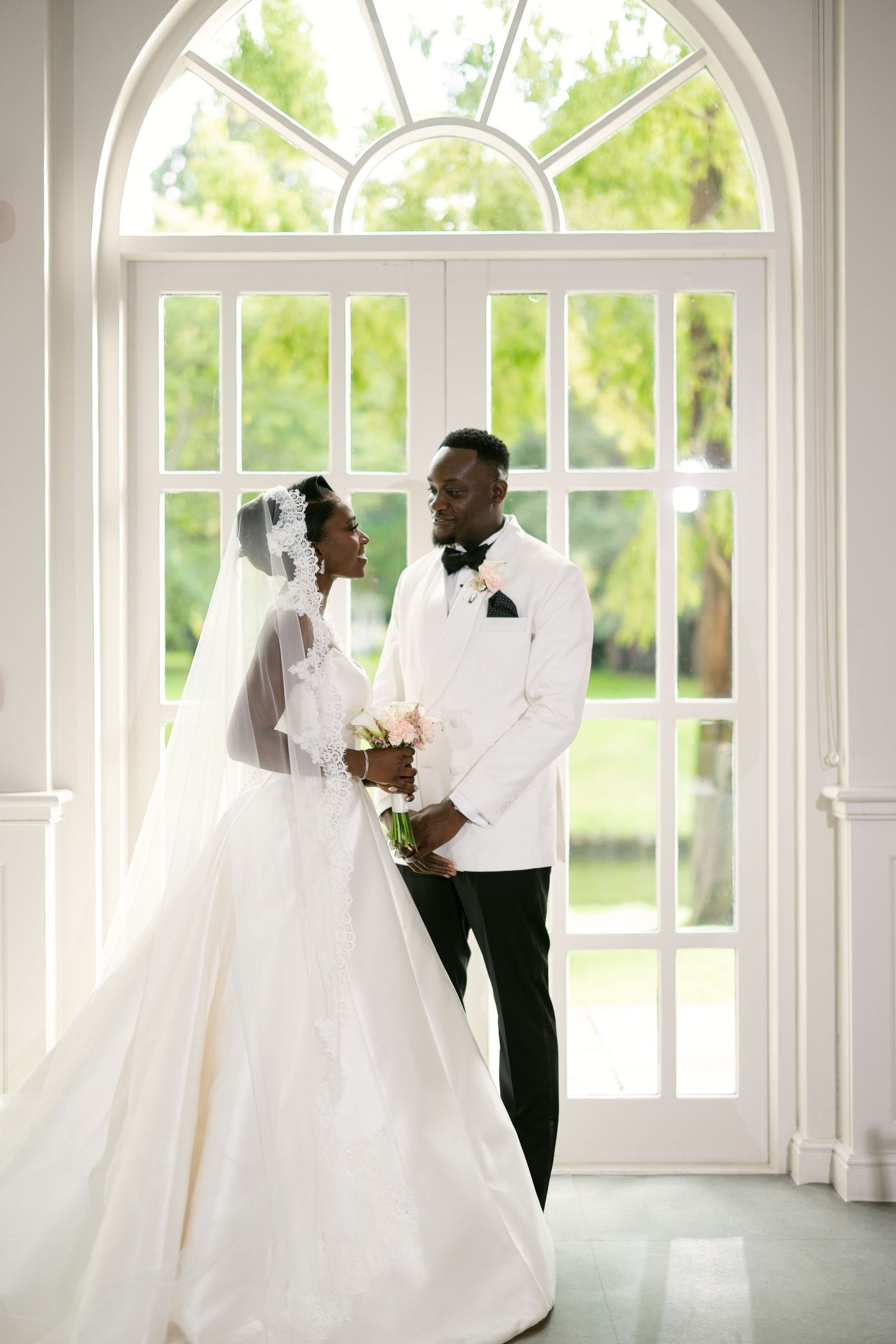 Black couple in wedding attire stand before large arched window, bride in white gown with veil, groom in white tuxedo jacket.