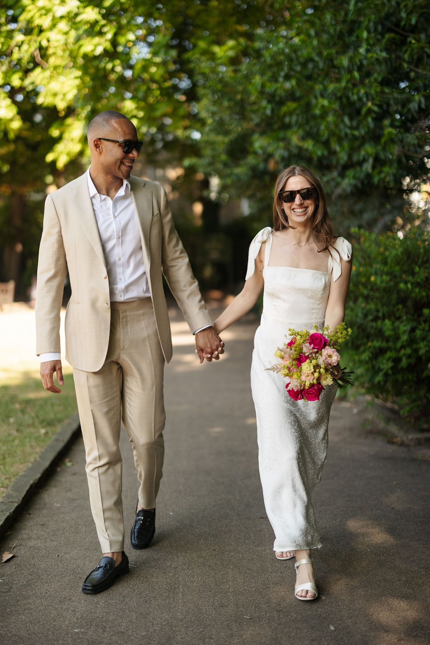 Smiling newlywed couple holding hands walking outdoors, bride in white dress with bouquet, groom in beige suit, both wearing sunglasses.
