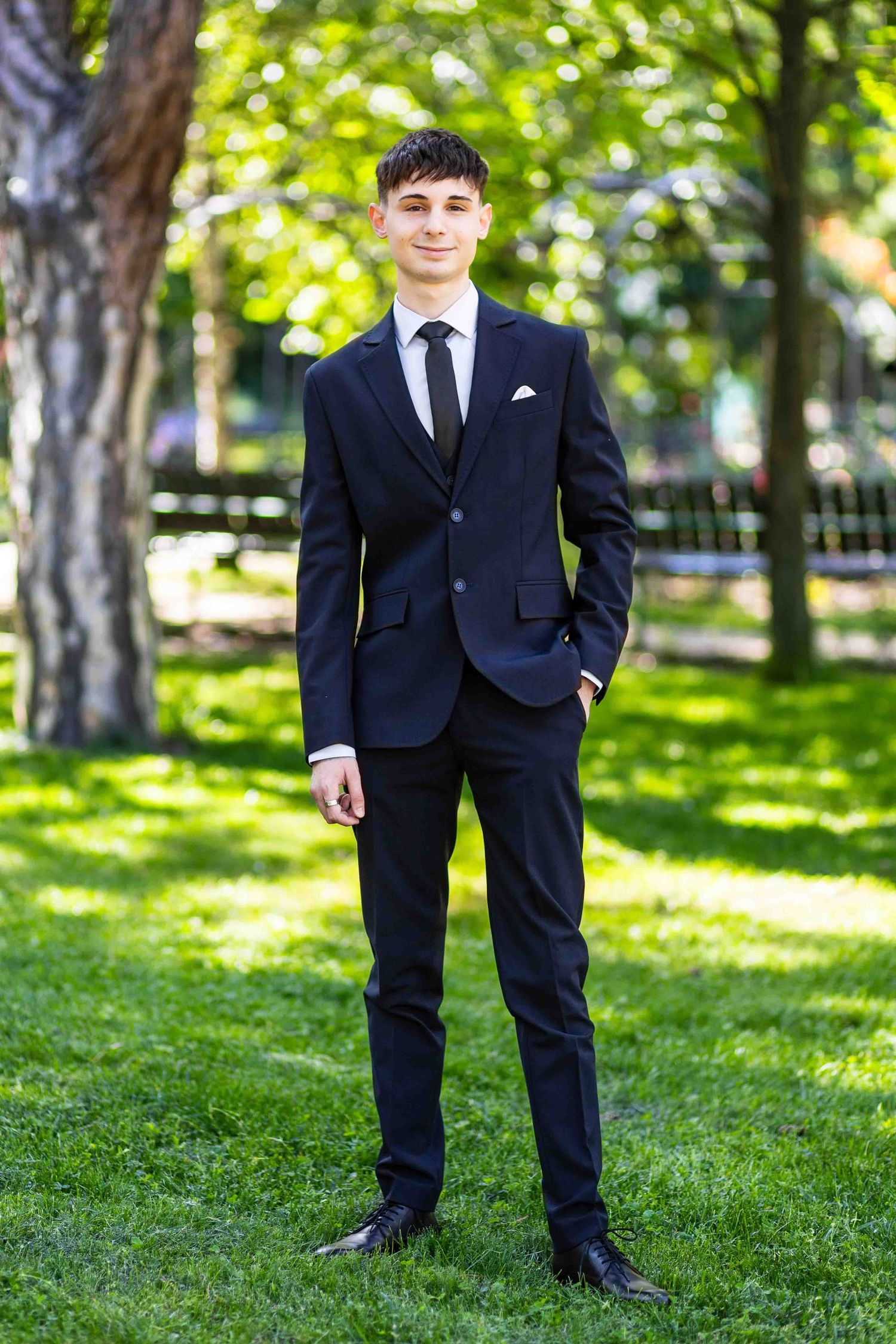 A young person in a black suit and tie poses in a park with trees and sunlit grass in the background.