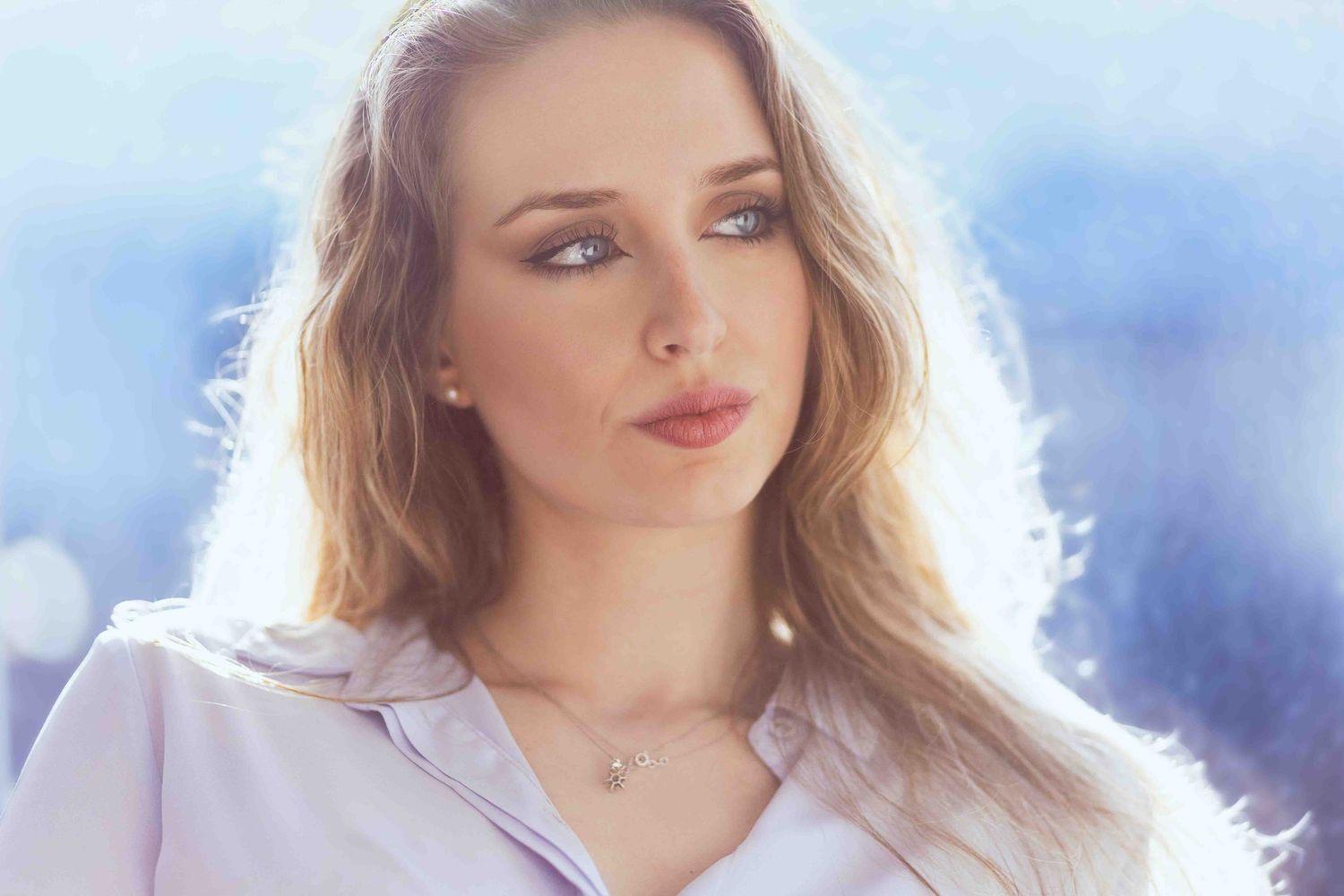 Portrait of a woman with long blonde hair wearing a white top against a soft blue background in natural lighting.