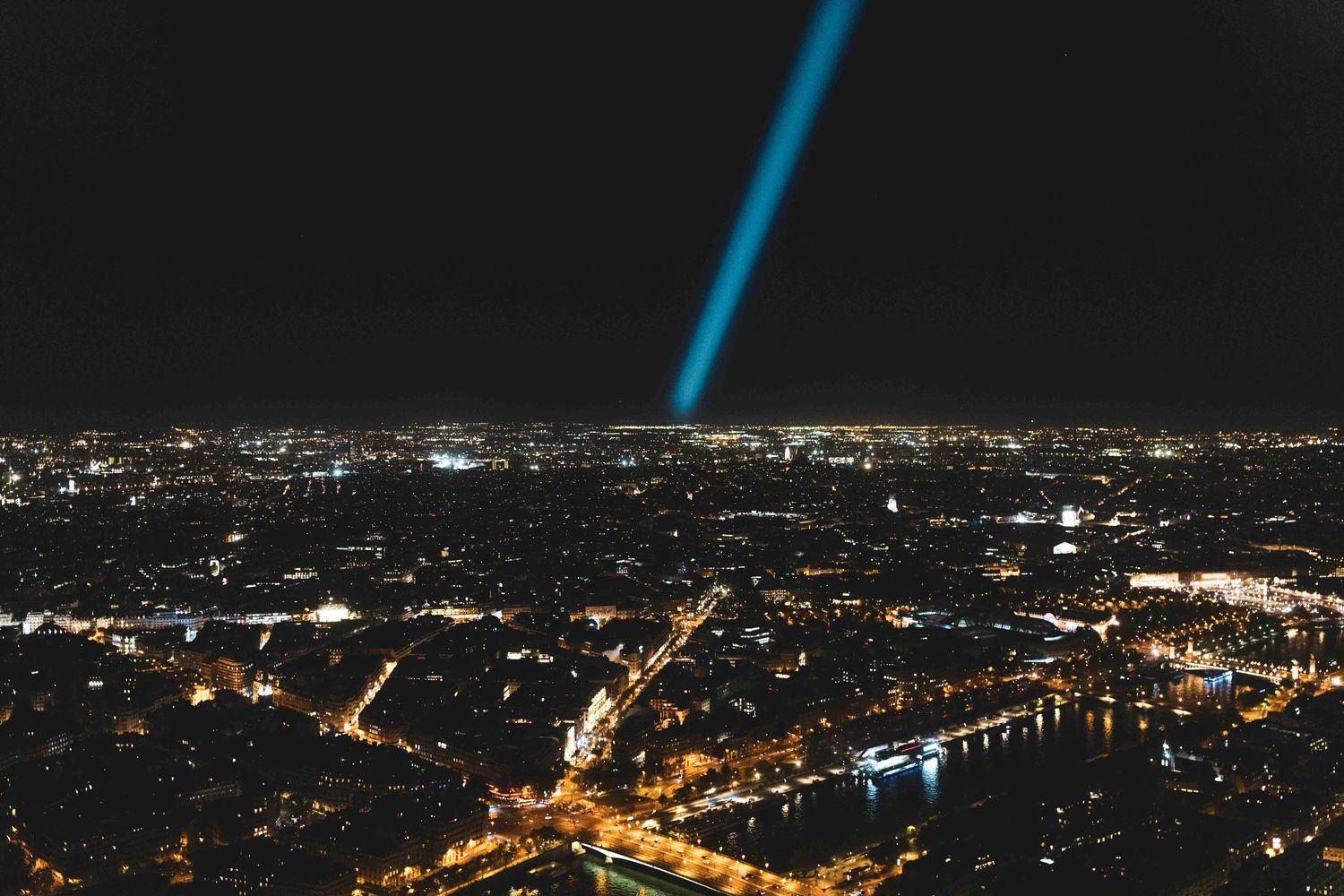 Bright blue beam of light pierces night sky above illuminated cityscape with winding river and glowing streets below.