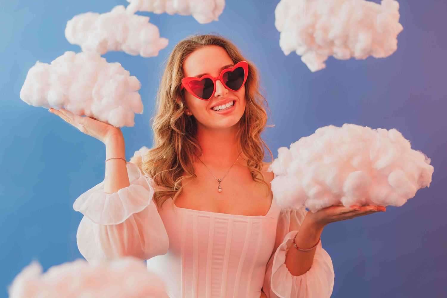Whimsical portrait with cotton cloud props against blue backdrop wearing red heart sunglasses.
