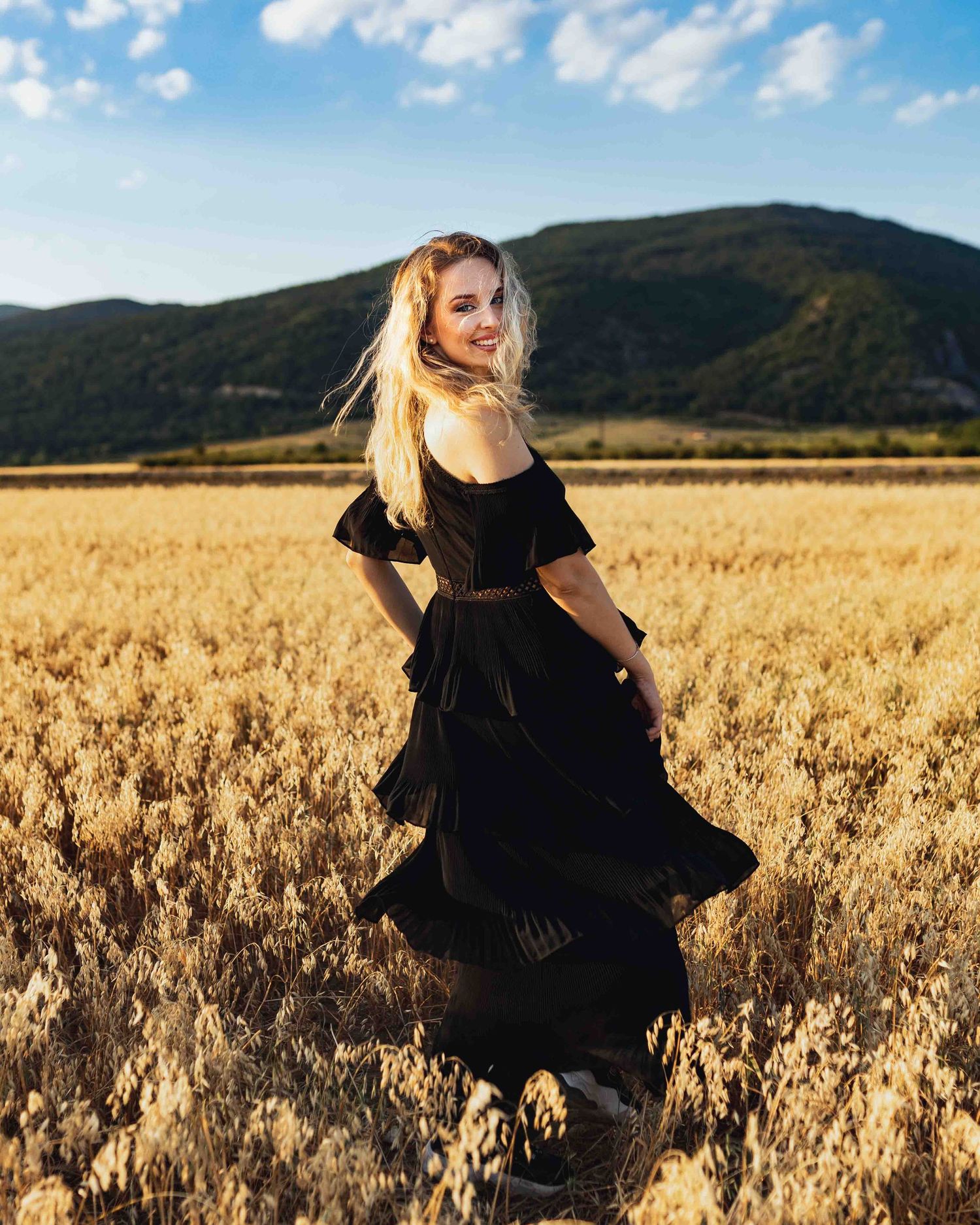A flowing black dress twirls in a golden wheat field against a backdrop of mountains and blue sky.