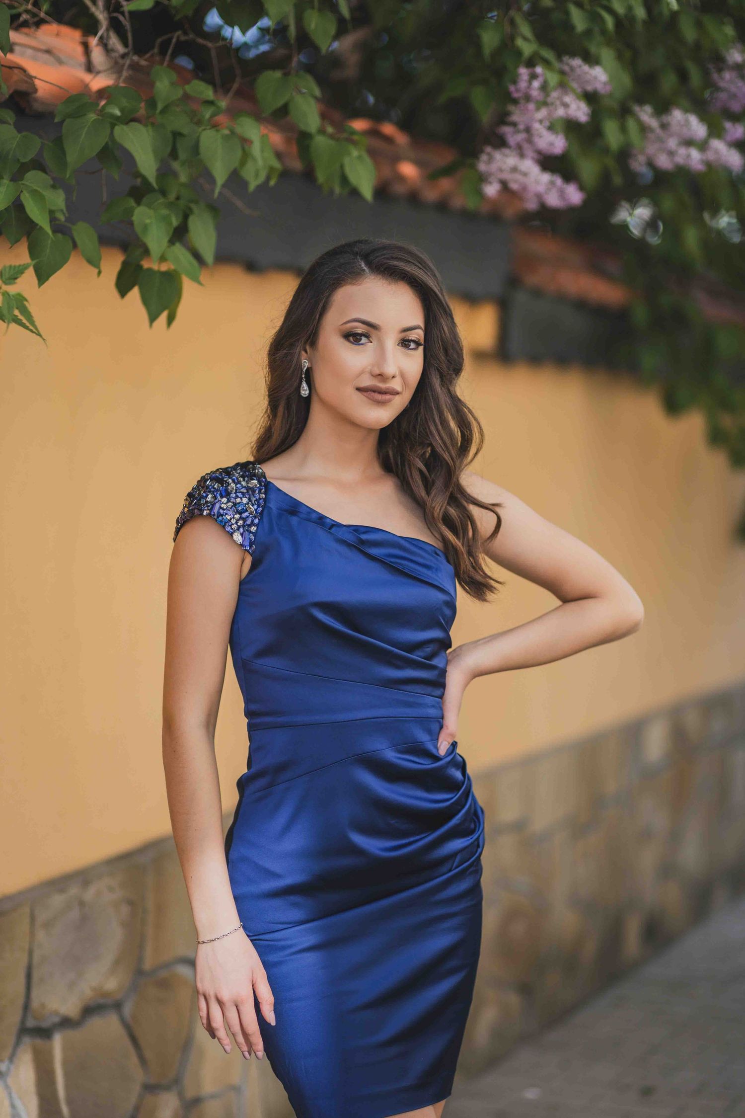 Elegant jewel-toned royal blue satin cocktail dress with ruched detailing and embellished cap sleeves against yellow wall.