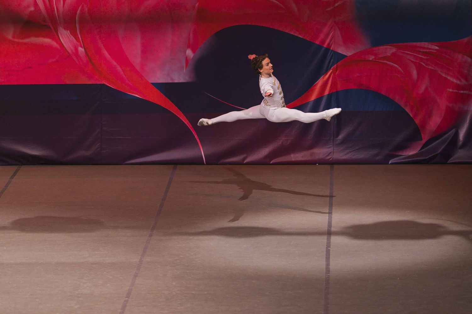 Professional ballet dancer in a red costume performs an elegant leap against a dark backdrop.