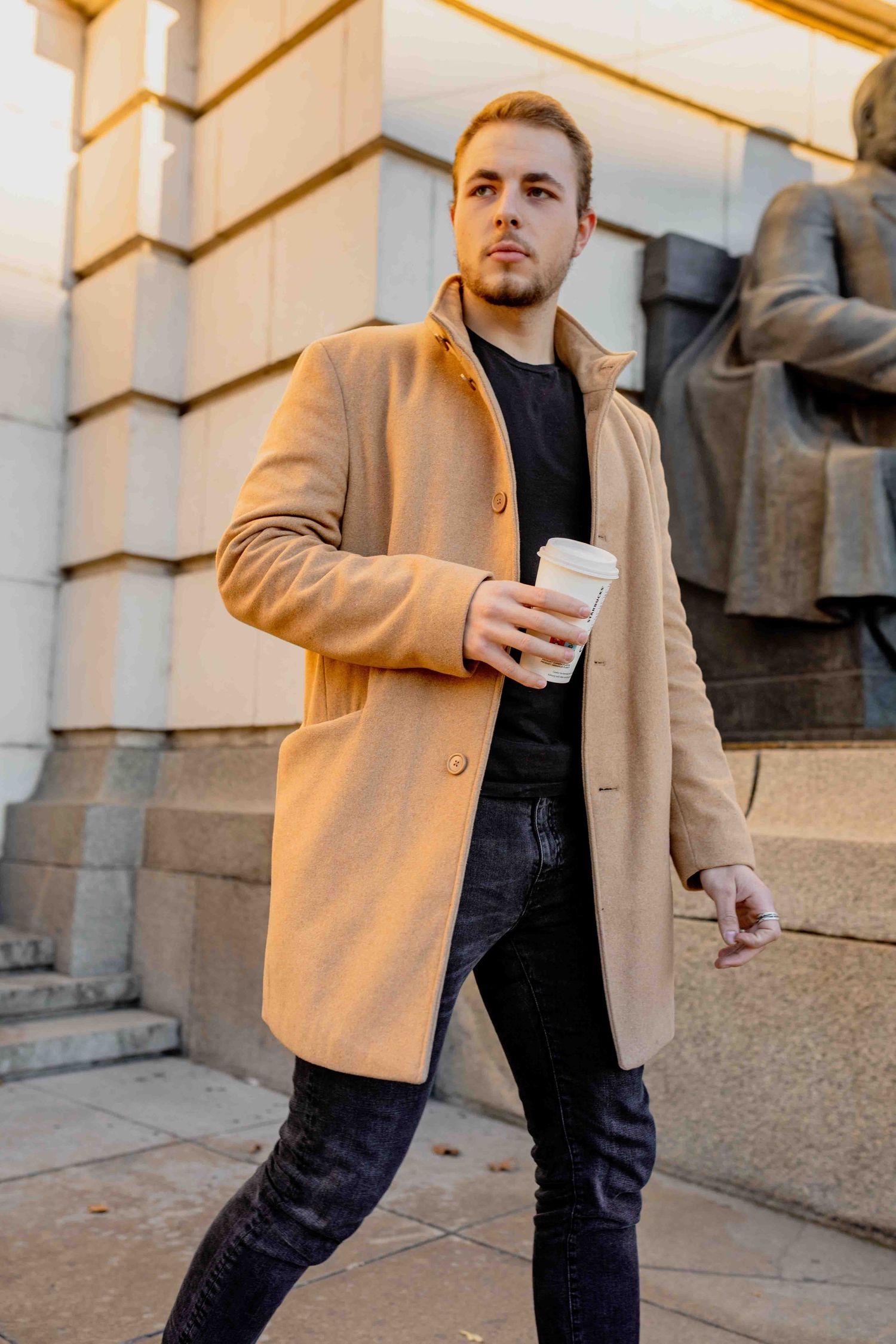 Stylish person in a camel coat holding coffee while walking past architectural columns on city street.