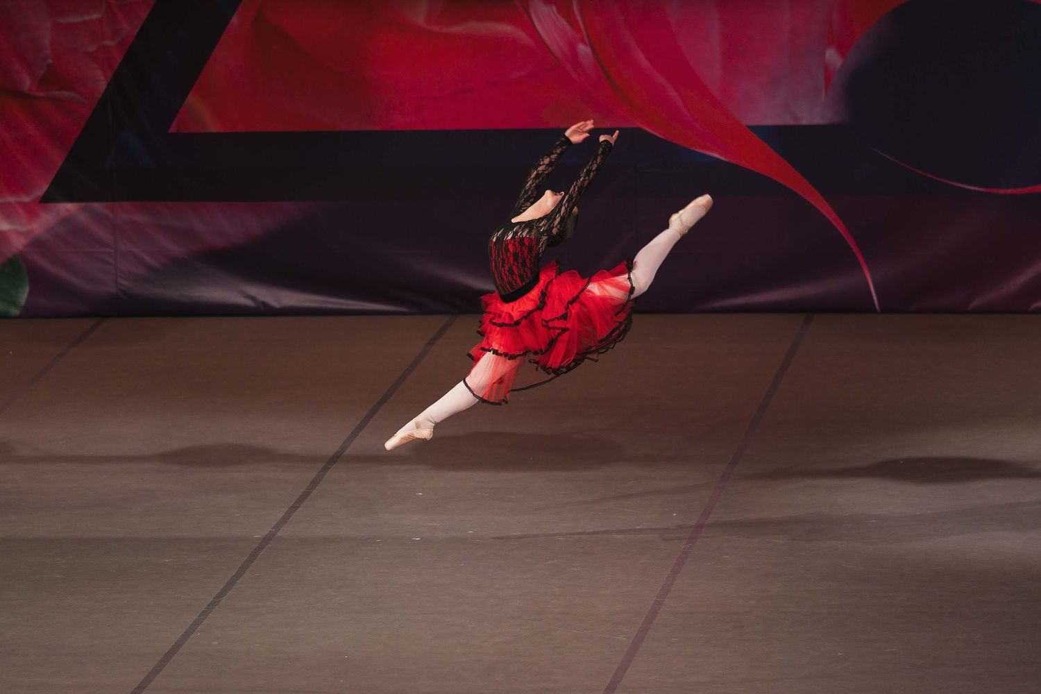 A ballet dancer in a red costume performs a dramatic leap across a stage during a performance.
