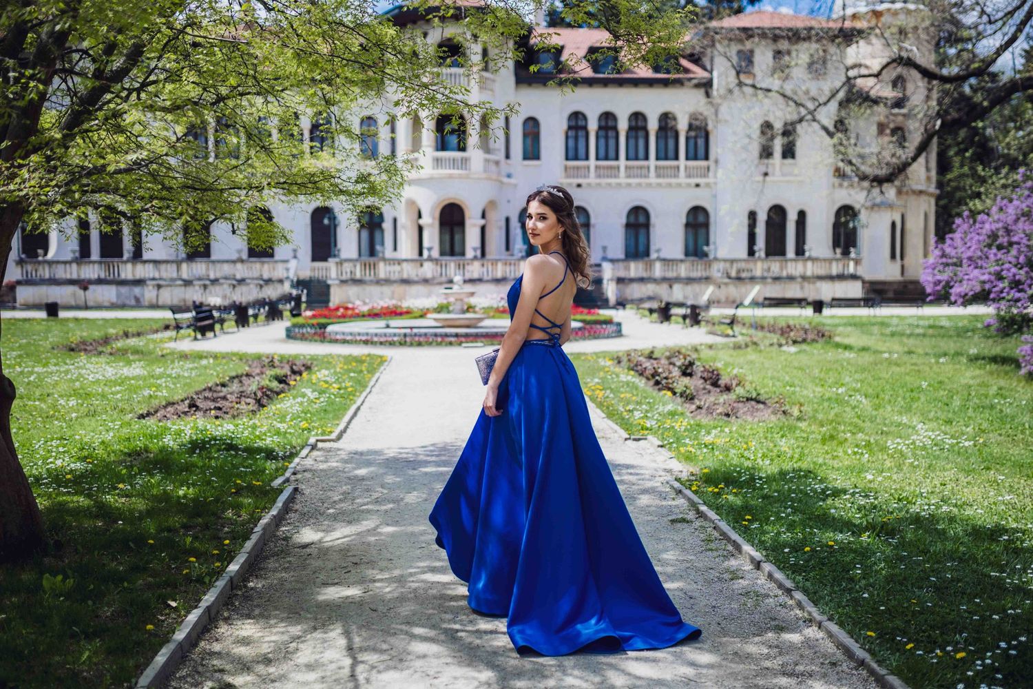 A bright royal blue formal gown is showcased against the backdrop of a white classical mansion with arched architecture.
