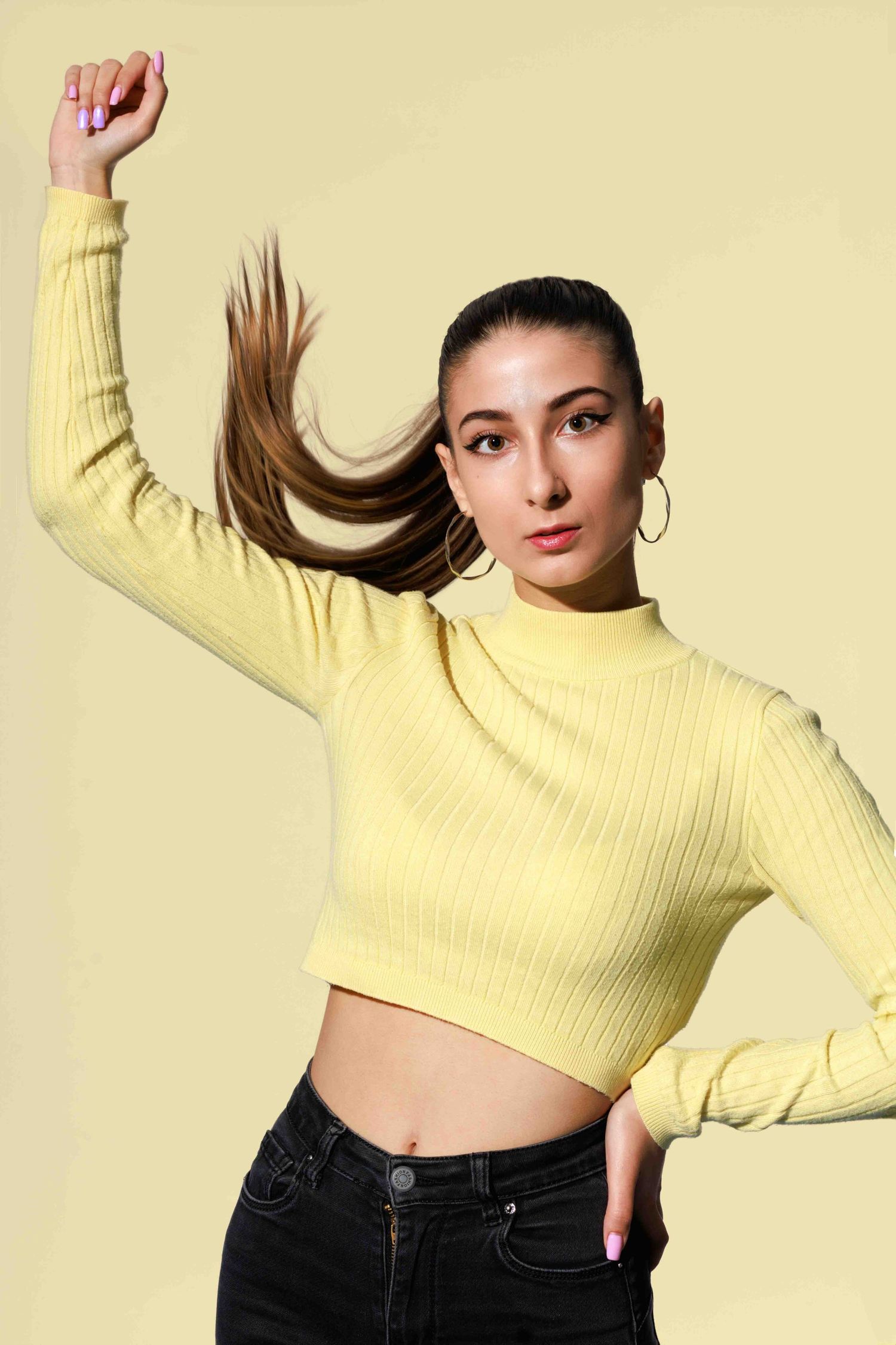Woman in yellow turtleneck crop top dancing with raised arm against light background.