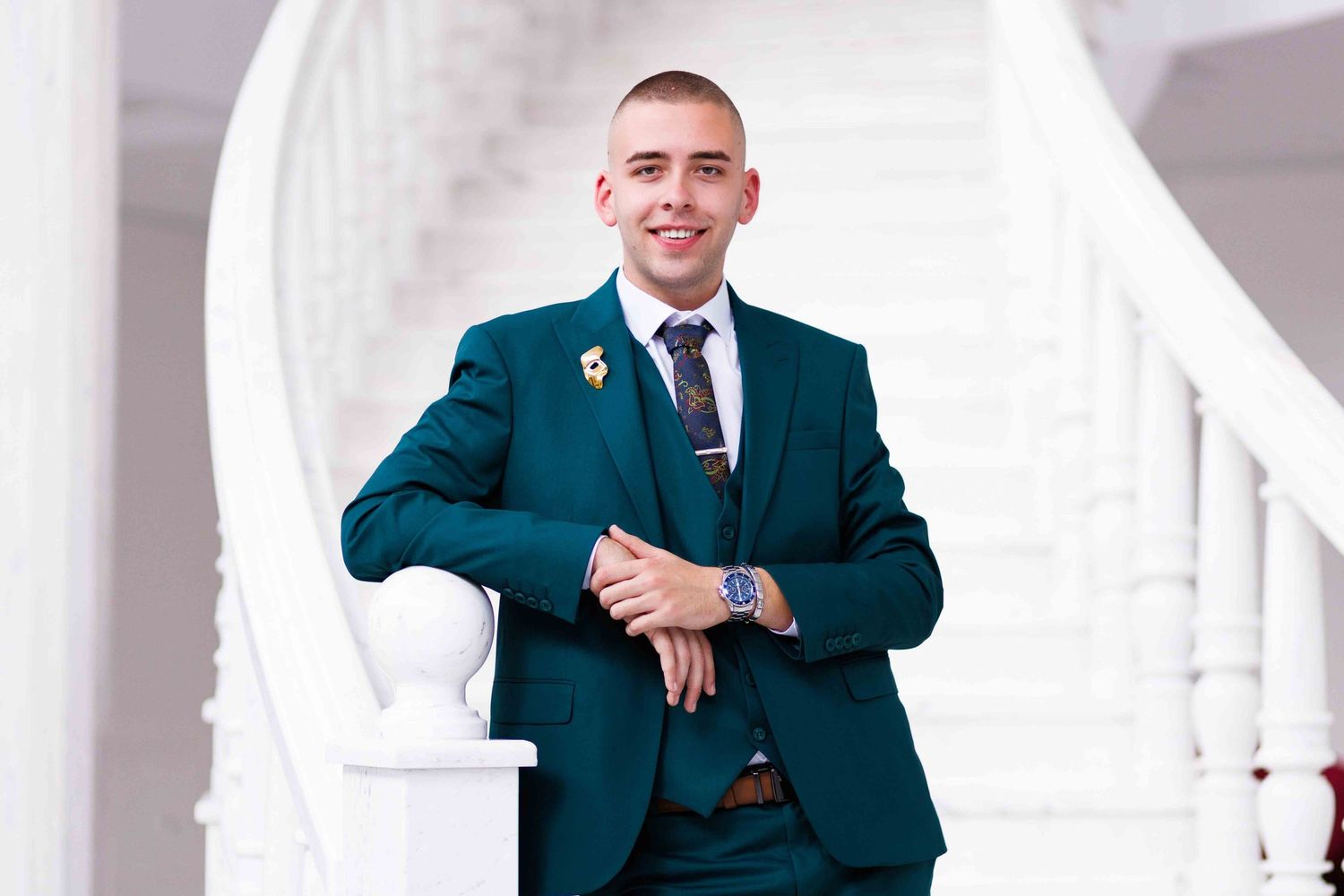 Professional portrait of someone in a teal suit and tie posing against a white architectural background.