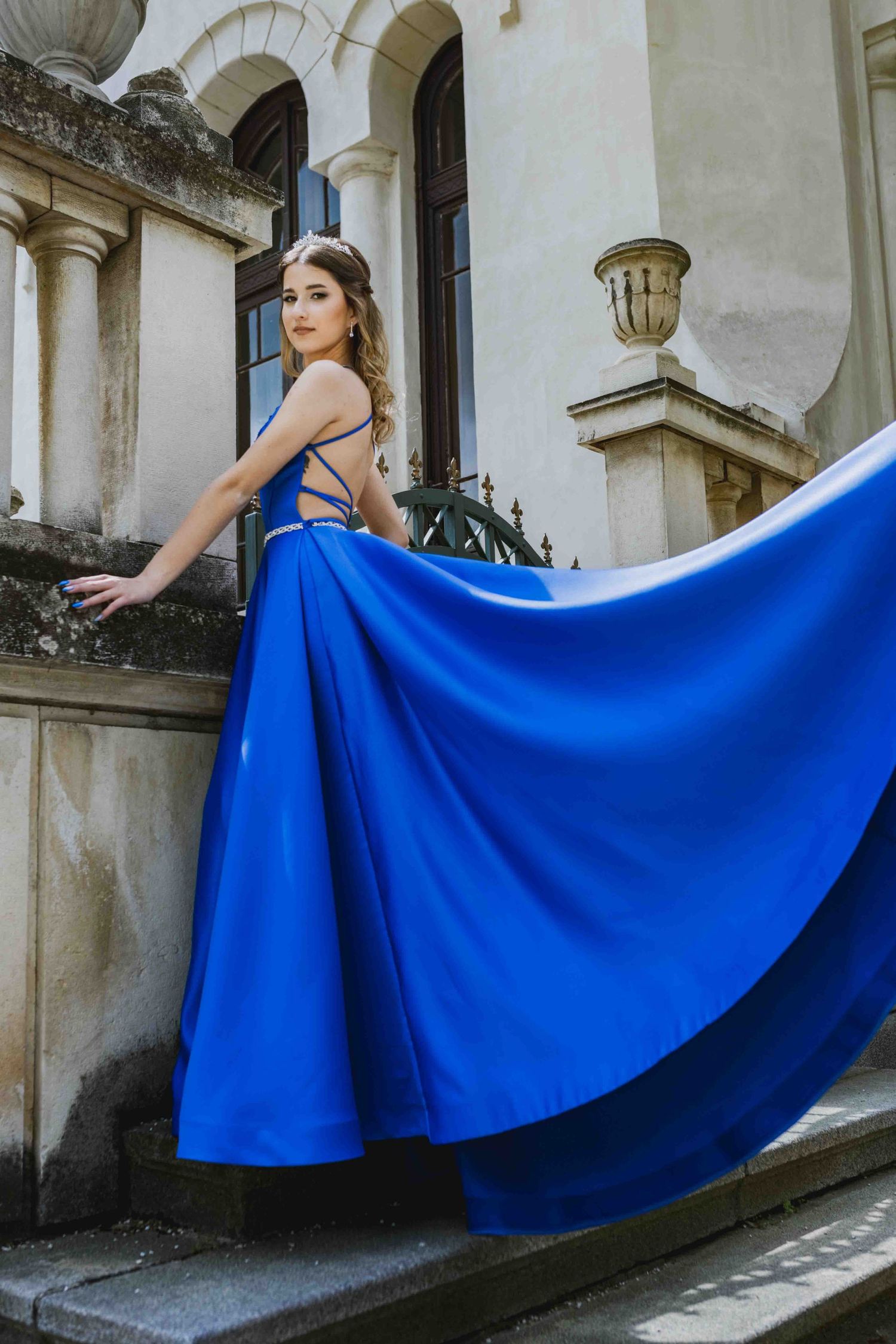 Elegant model in a flowing royal blue evening gown poses on stone steps against classical architecture.