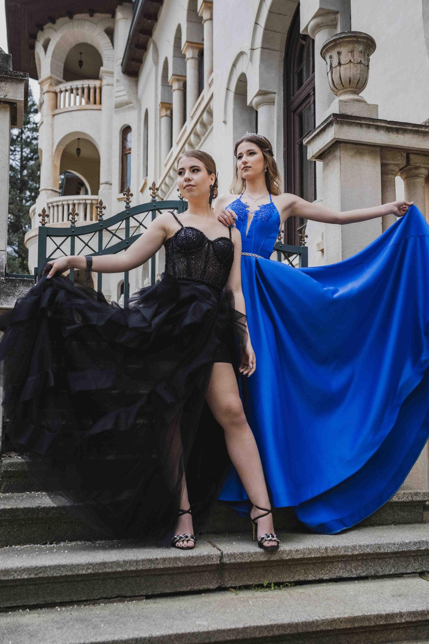 Two models pose dramatically on grand mansion steps wearing elegant black and royal blue gowns with flowing skirts.