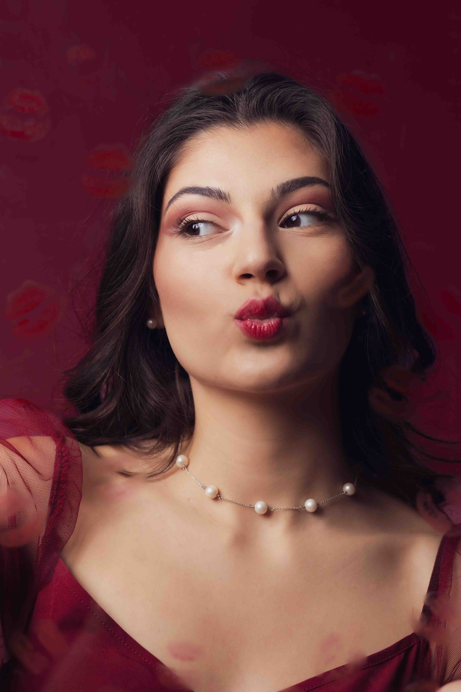Portrait against red background with pearl necklace showing playful expression and wearing red dress.