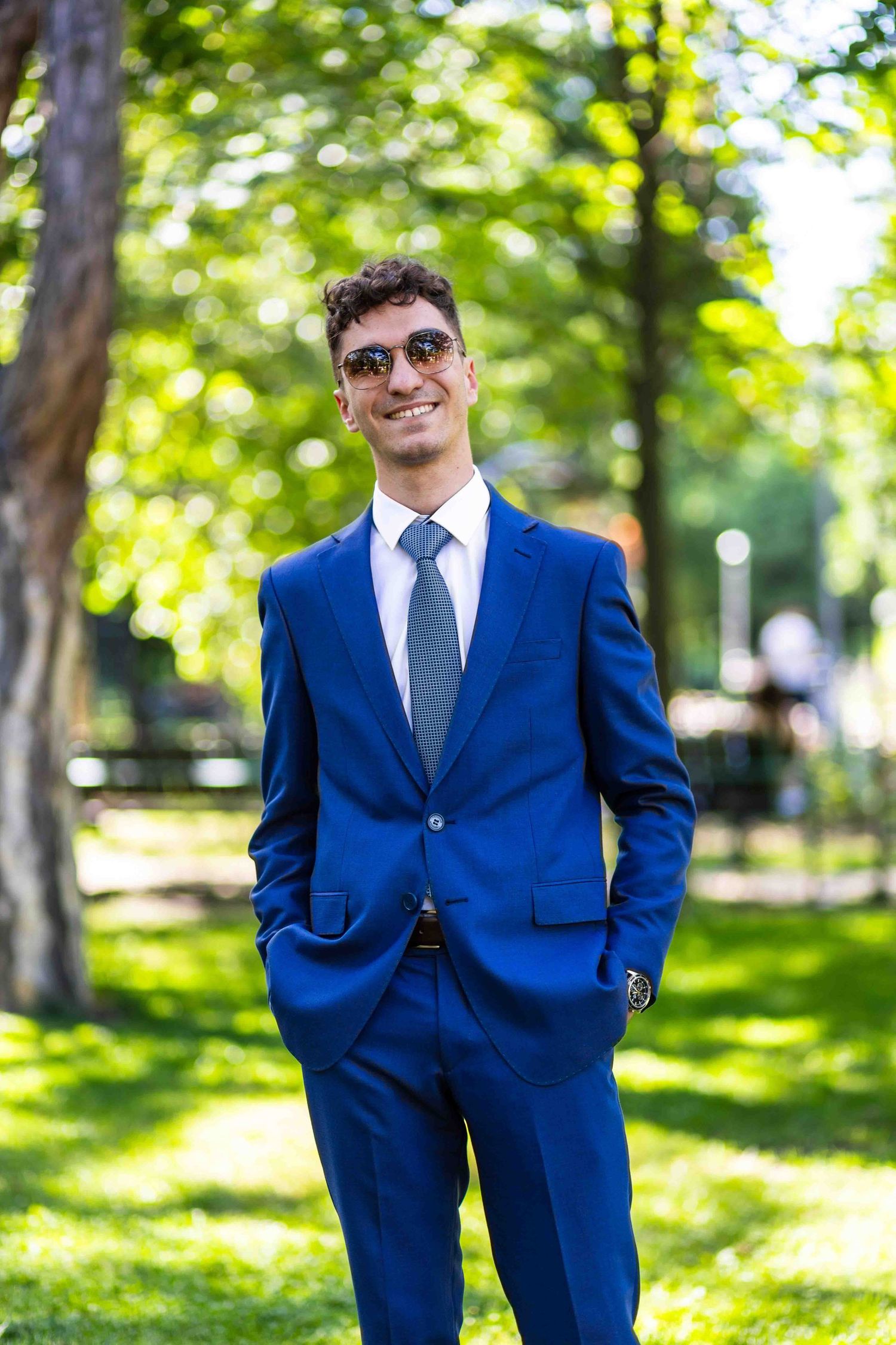 Professional portrait of person in navy blue suit standing in sunny park with blurred green background.