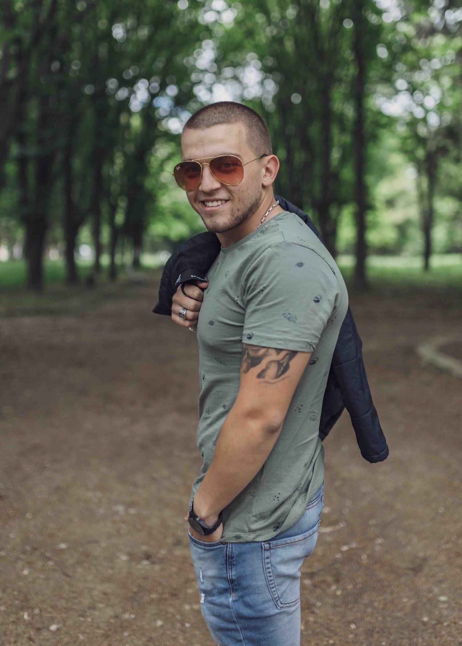 Someone wearing sunglasses and a sage green t-shirt poses casually in a wooded park setting.