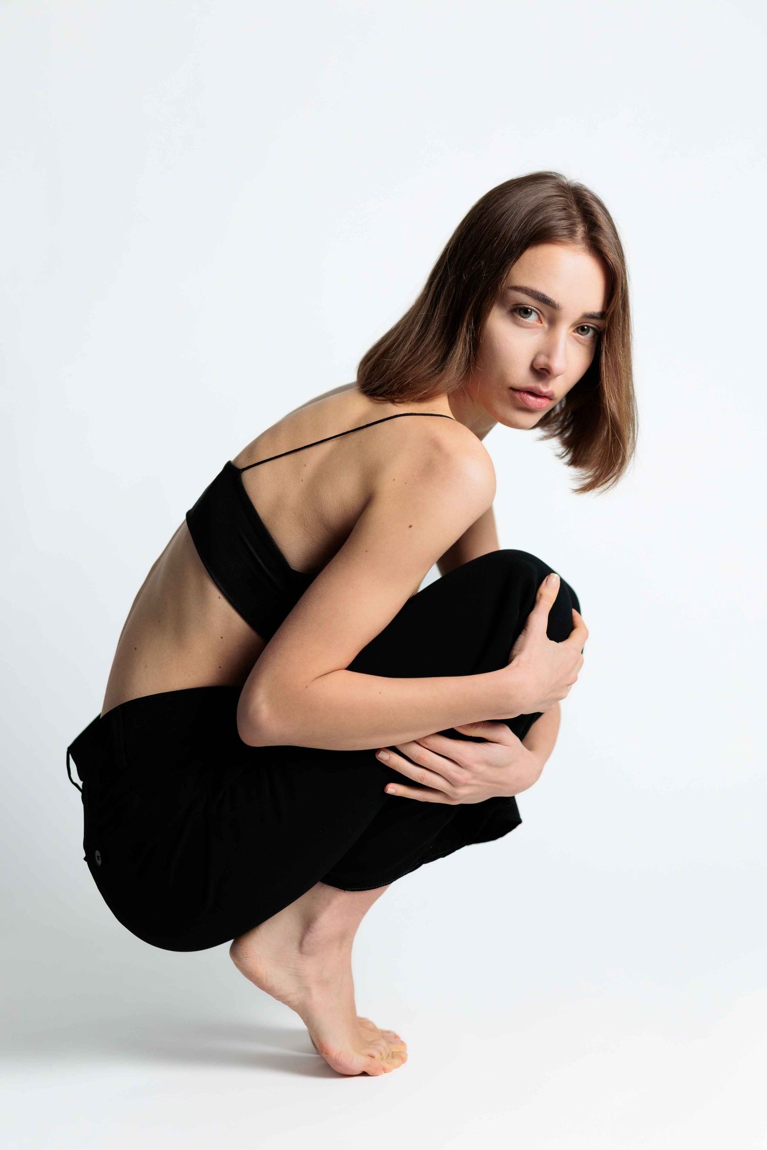 A model in black athletic wear crouches low on a white background in a minimalist artistic pose.