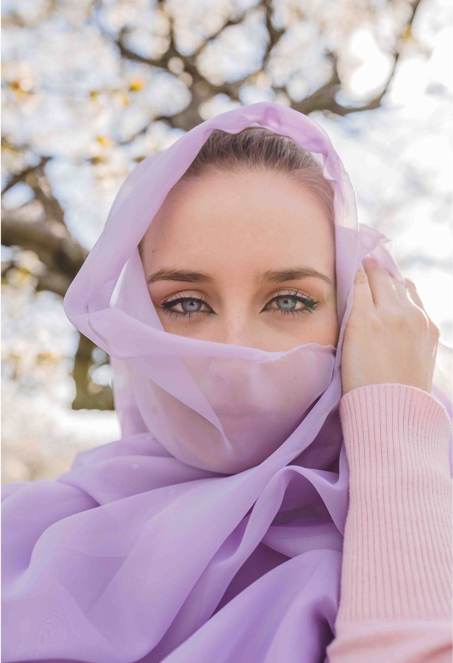 Eyes peer mysteriously through a flowing lilac veil against a soft spring background.