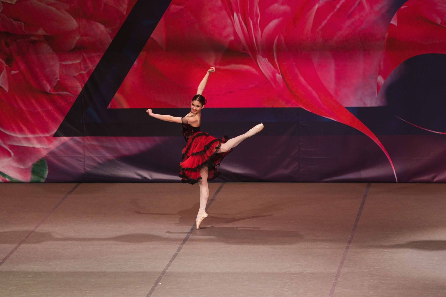 A ballet dancer in a flowing red dress performs a graceful split leap against a dramatic crimson background.
