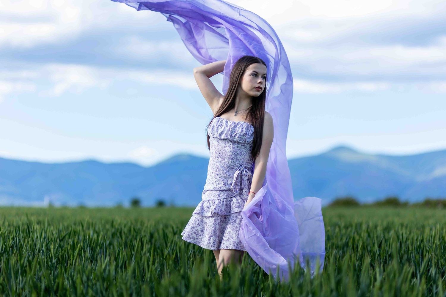 A purple flowing fabric billows in the wind above a green field with mountains in the background.