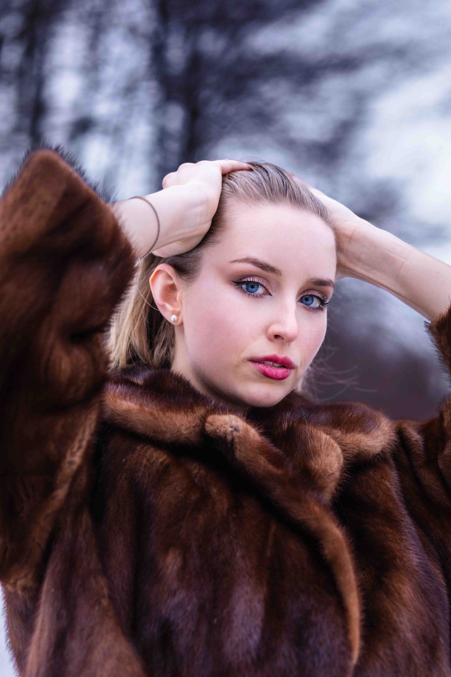 A woman wearing a luxurious brown fur coat poses outdoors in winter with light makeup and windswept hair.