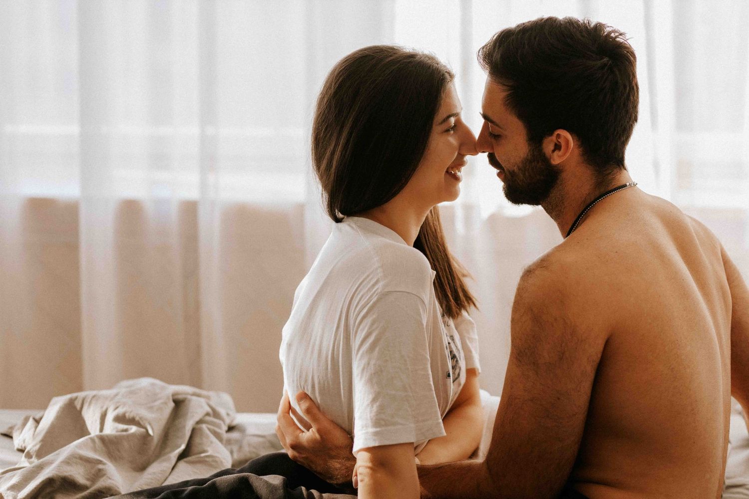Intimate couple sharing a tender romantic moment while sitting together in a bright sunlit bedroom.