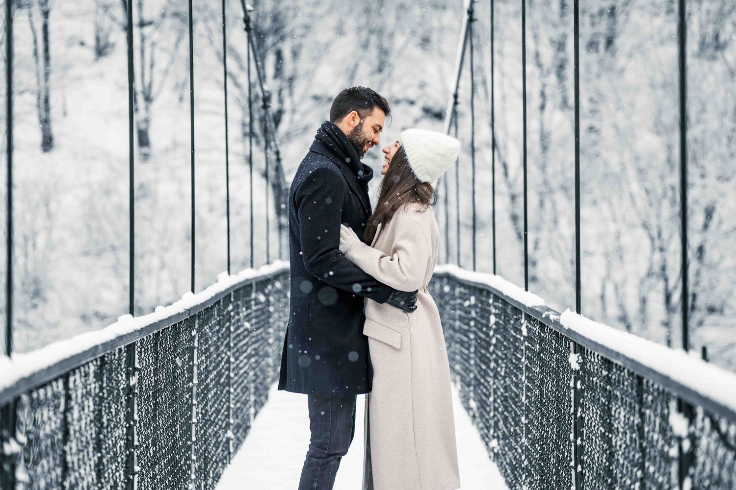 A romantic couple embraces on a snow-covered bridge in winter with frost-covered trees in the background.