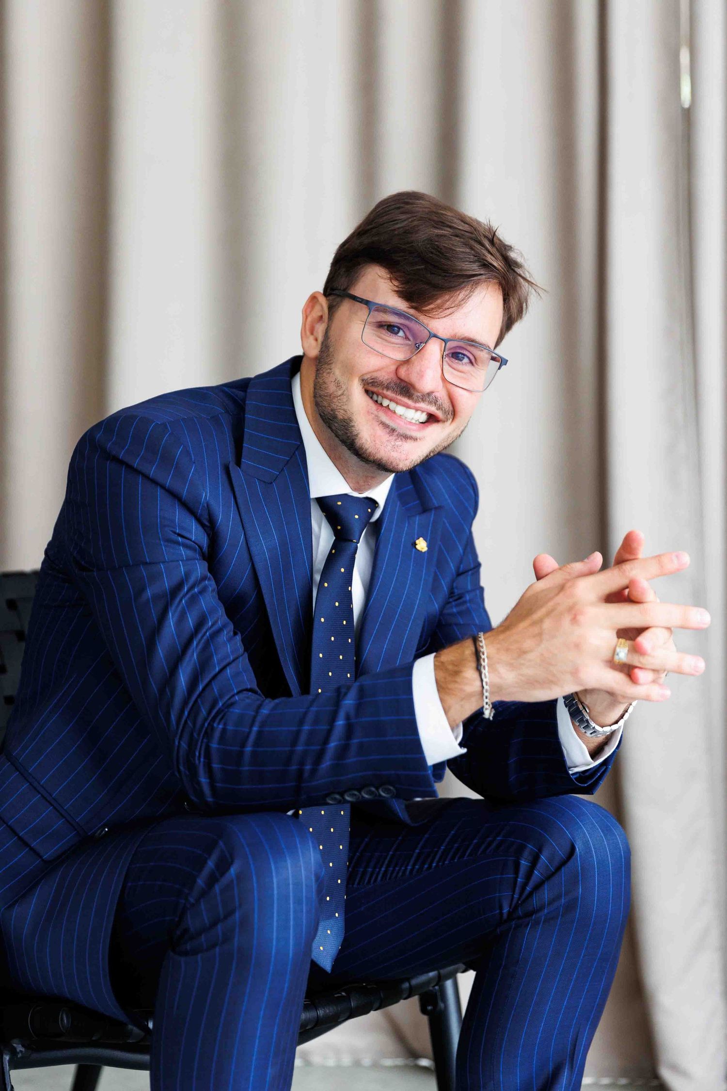 Professional wearing a navy pinstriped suit, blue tie and glasses sits in relaxed pose against light curtain background.
