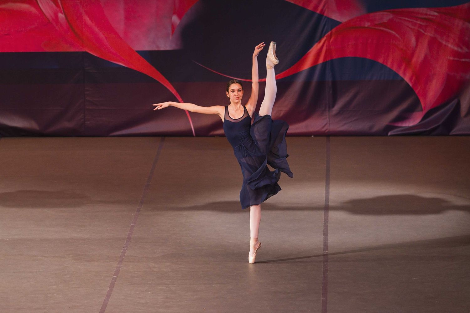 A ballet dancer performs an elegant arabesque pose on stage with a dramatic red and black backdrop.