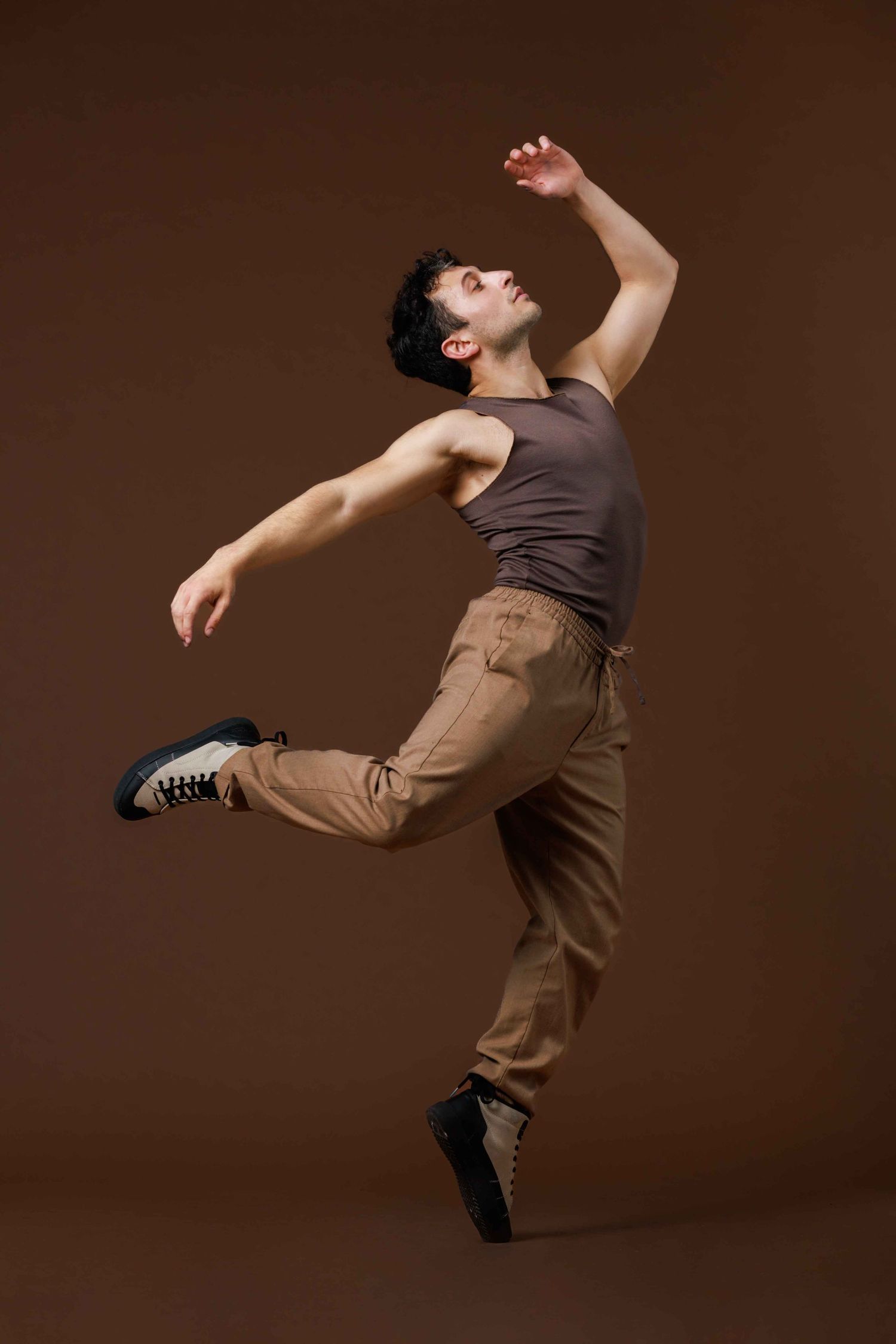 A contemporary dancer in a brown tank top and khaki pants performs a dramatic airborne leap movement on a dark background.