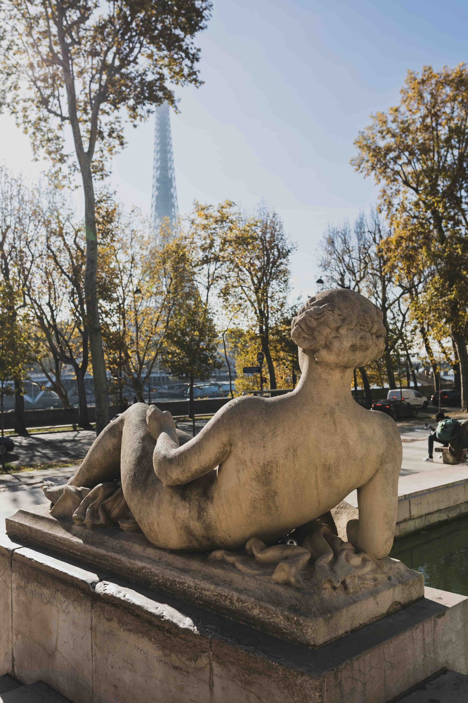 A classical stone statue reclining near the Eiffel Tower in Paris with autumn trees in the background.