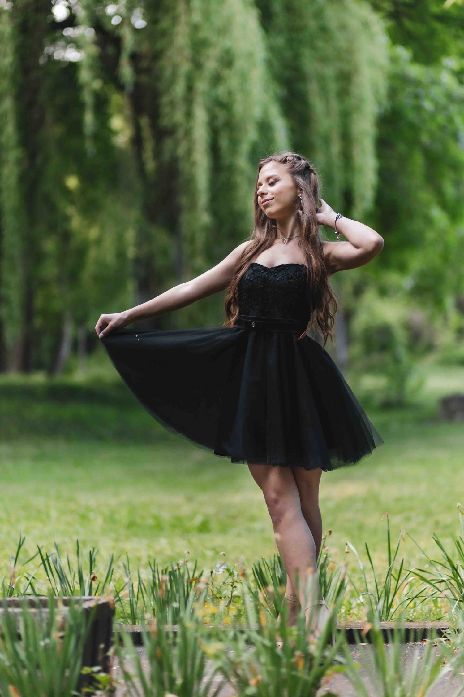 A graceful dancer in a black tulle dress poses elegantly in a lush garden setting with weeping willows in the background.