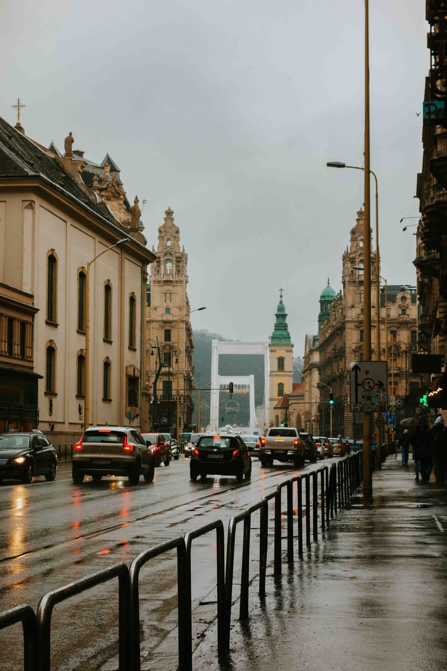 European city street lined with historic buildings on a rainy day, with wet cobblestones and car headlights reflecting in puddles.