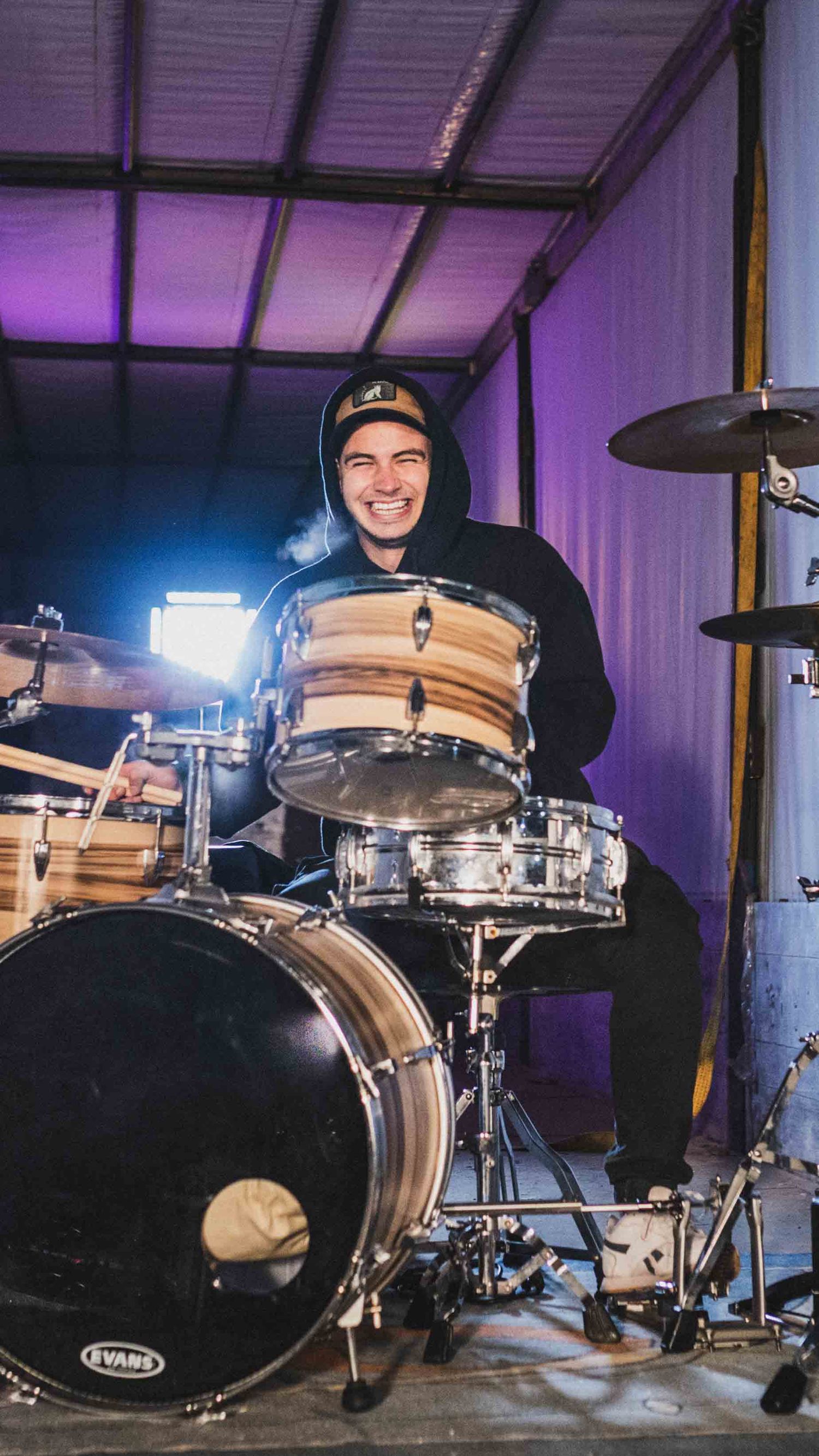 A drummer in a black hoodie smiles while sitting behind a wooden drum kit in a purple-lit room.