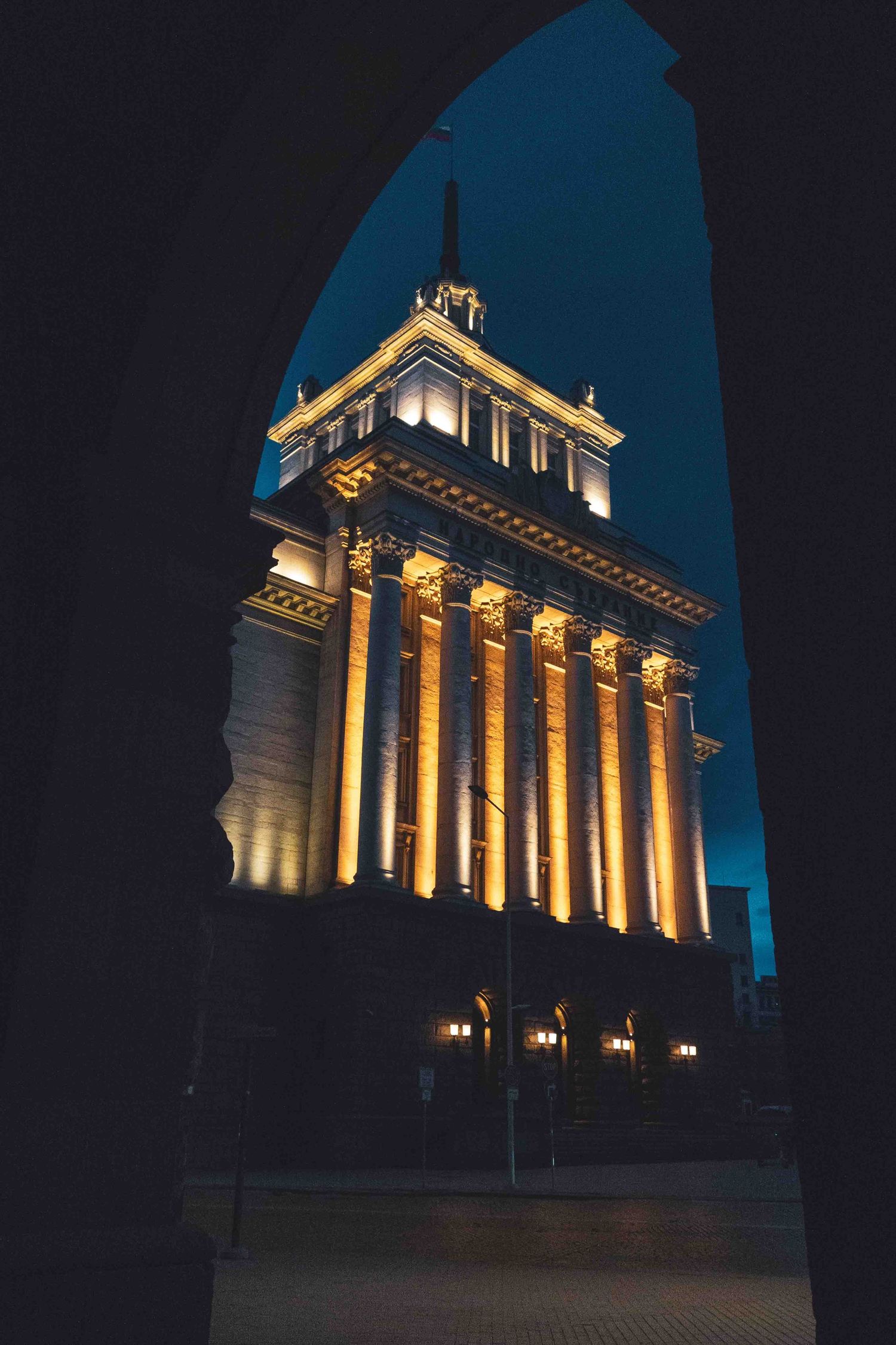 Illuminated neoclassical building with tall columns viewed through an archway at night against a dark blue sky.