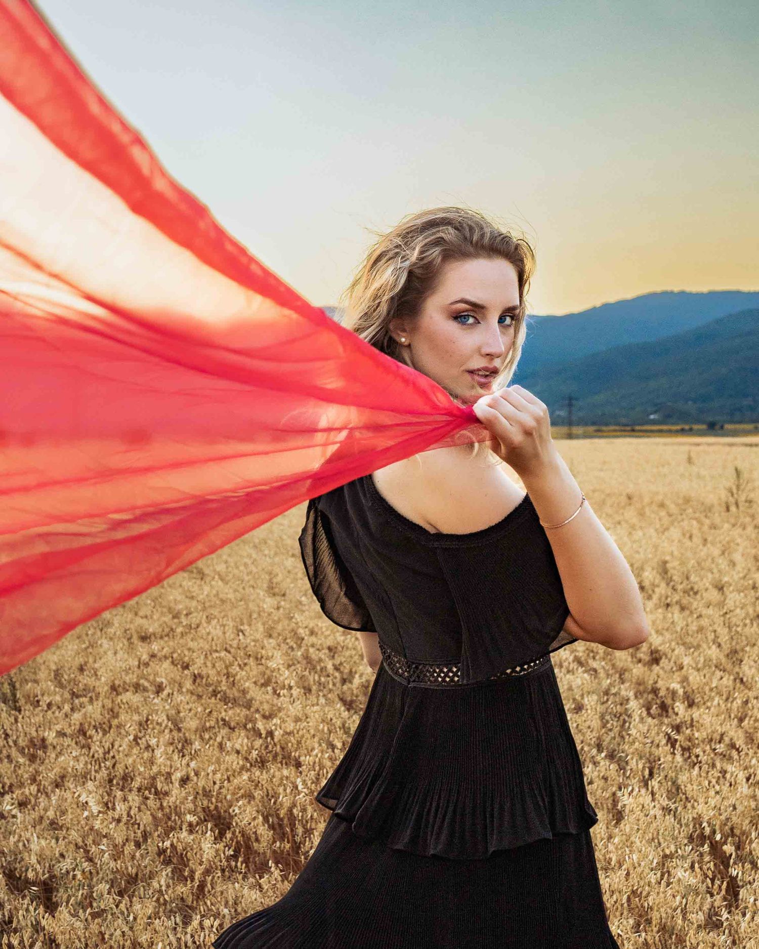 A person in a black dress stands in a wheat field while red fabric flows dramatically in the breeze at sunset.