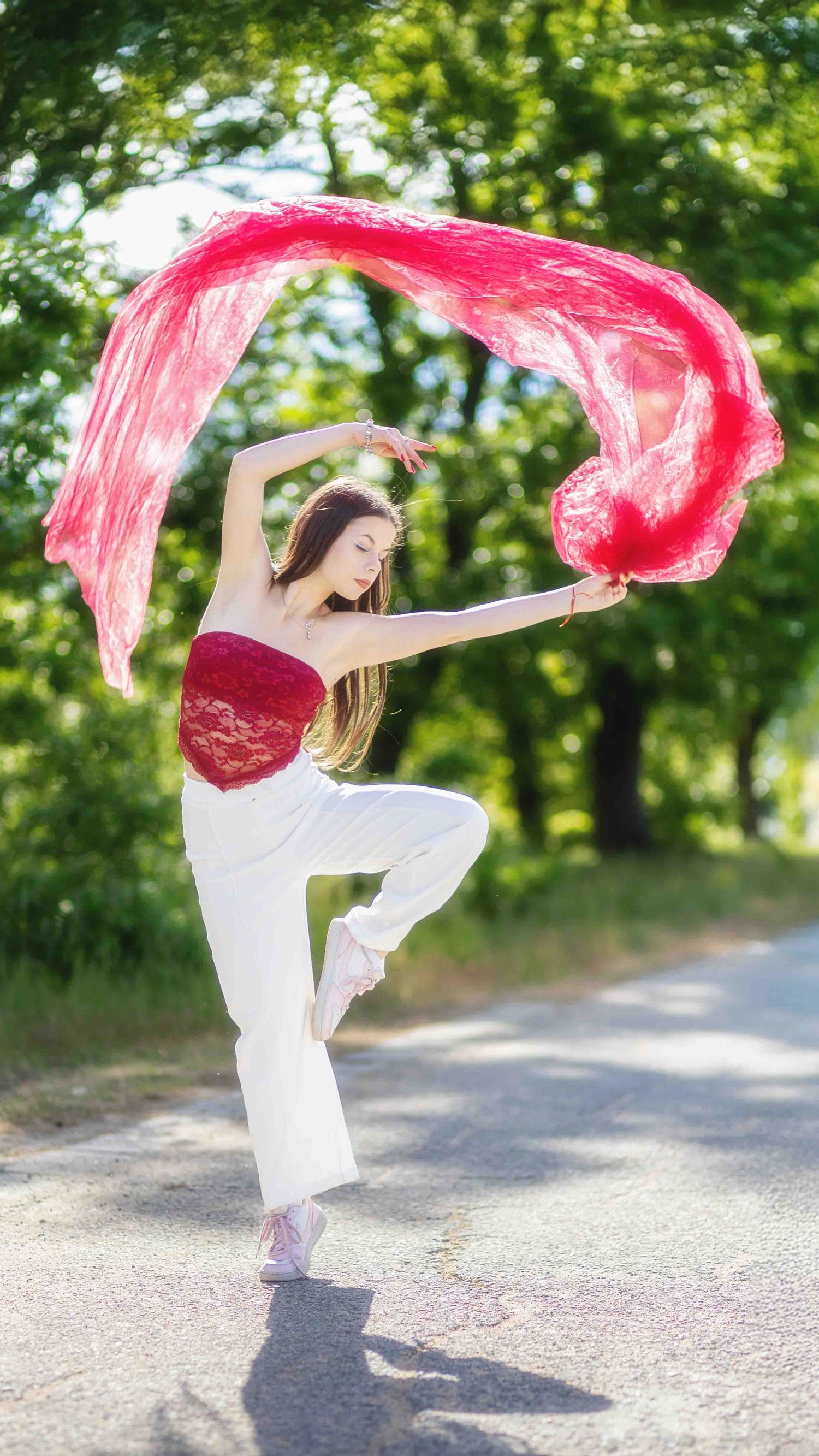 A graceful dancer in white pants and red top performs on a sunlit path while waving a flowing red silk scarf.