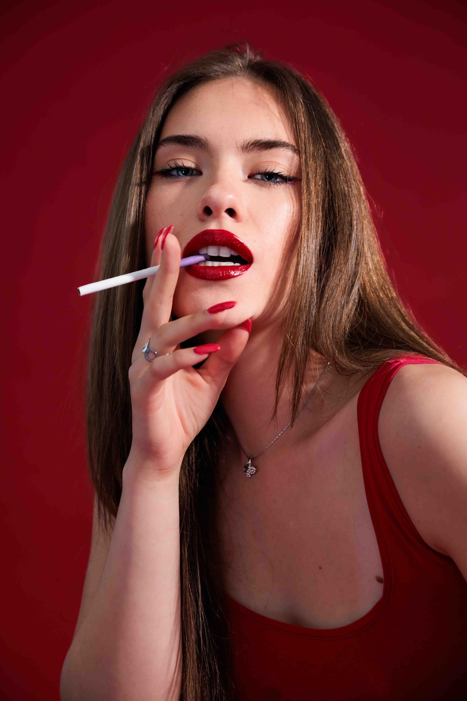 Portrait against a red background, showing long brown hair, red lipstick, and red nails, holding a cigarette.
