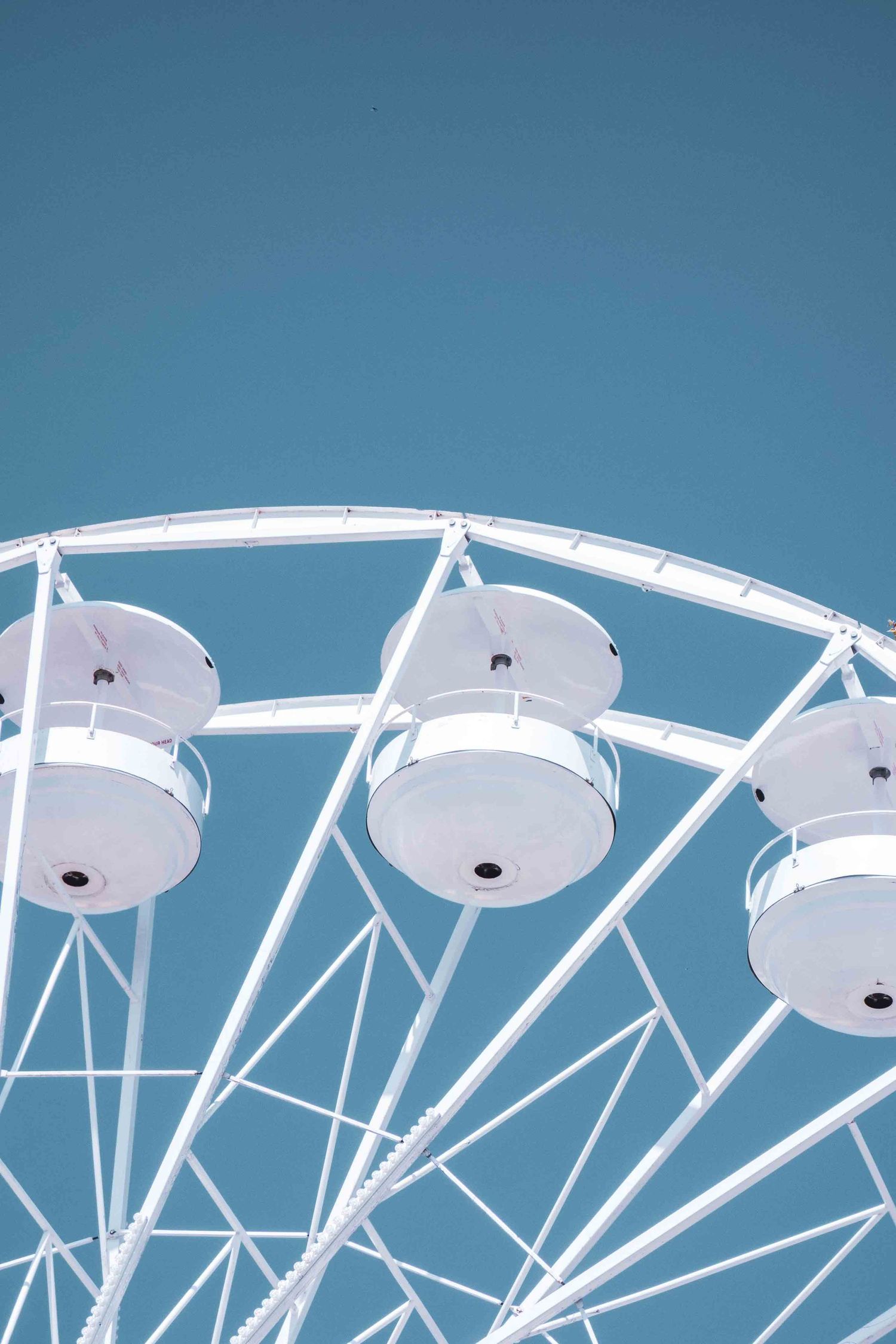 White enclosed gondolas on a Ferris wheel against a bright blue sky in a minimalist composition.