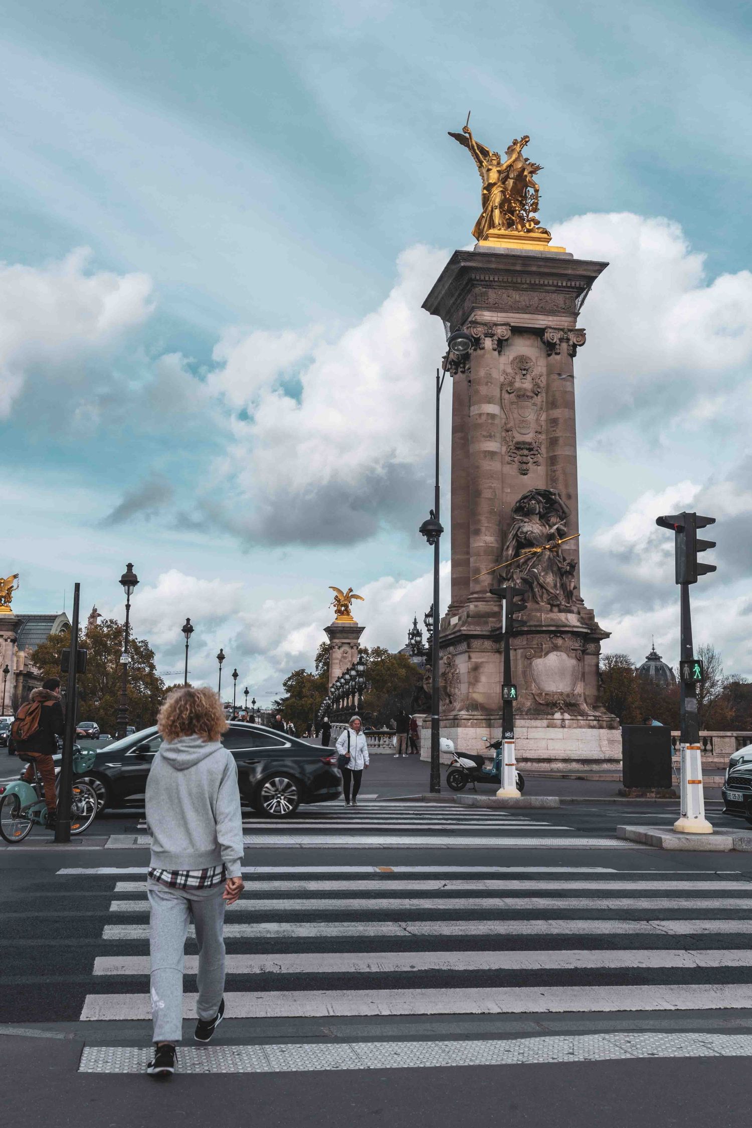 A person stands at a crosswalk near the iconic Pont Alexandre III in Paris with its golden winged statue atop a column.