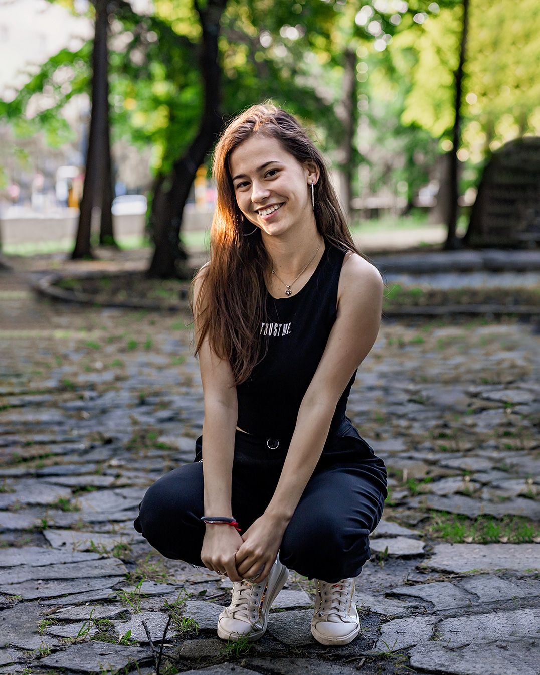 A smiling person in a black sleeveless top crouches on a stone path among trees in a park setting.