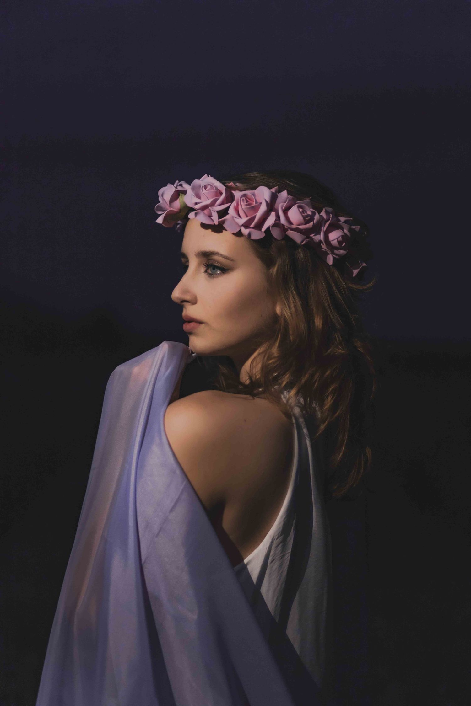 A figure wearing a pink floral crown and sheer veil poses against a dark background in an ethereal fashion portrait.