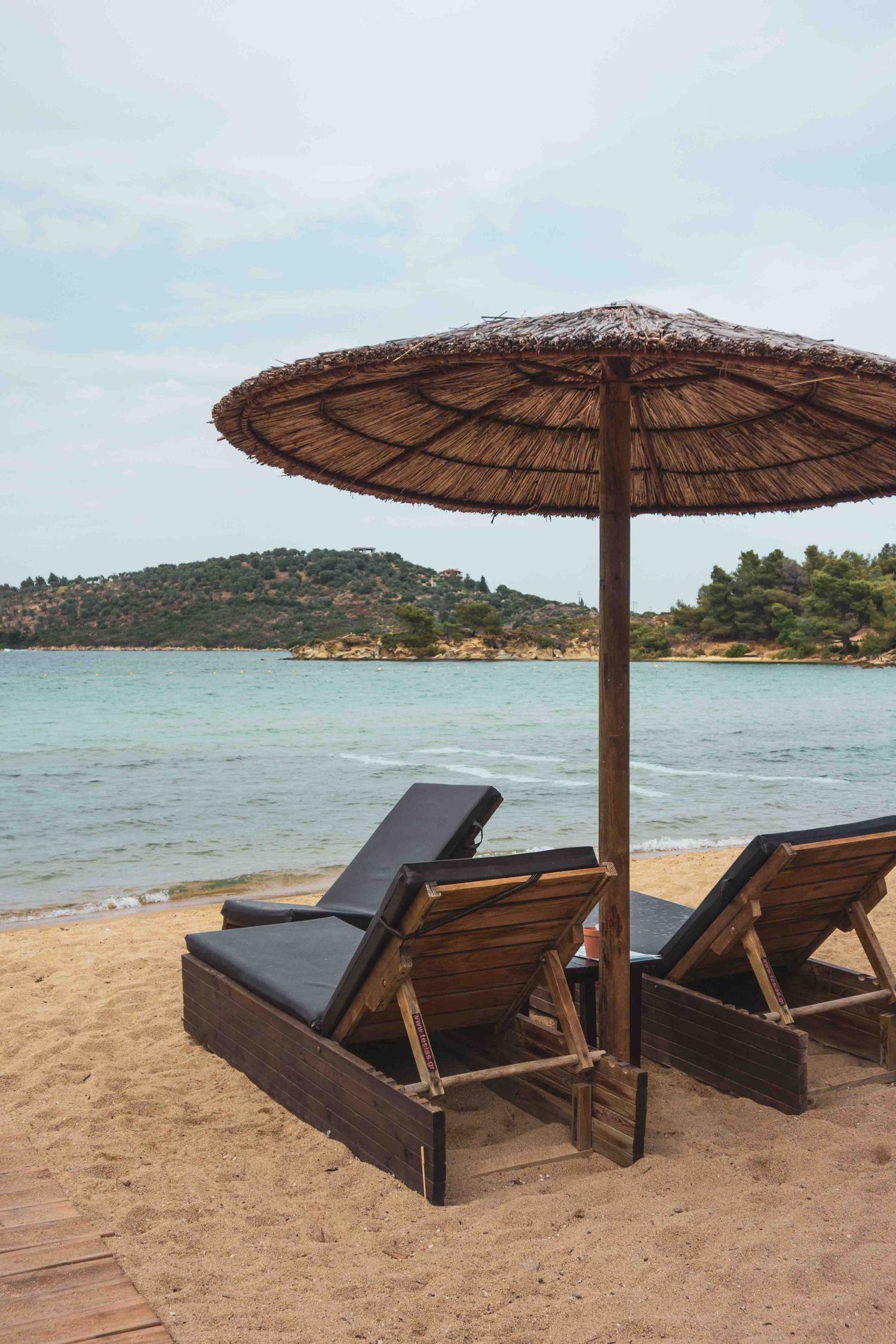 Beach lounge chairs with thatched umbrellas line a sandy beach overlooking calm tropical waters and distant hills.