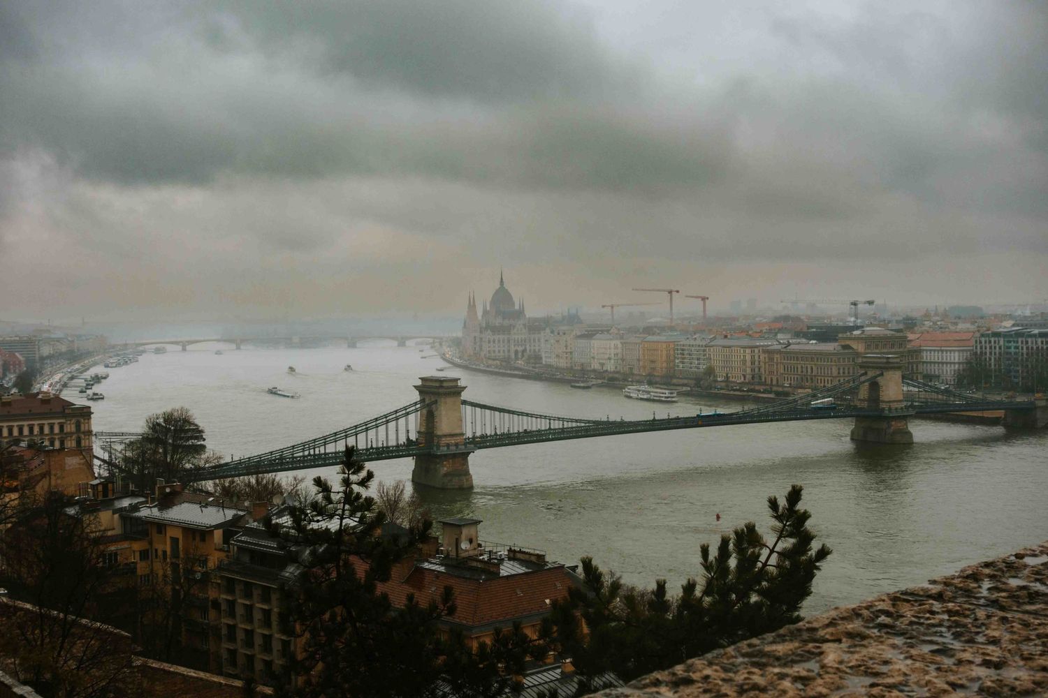Moody aerial view of Budapest's Chain Bridge and Parliament Building along the Danube River under stormy gray skies.