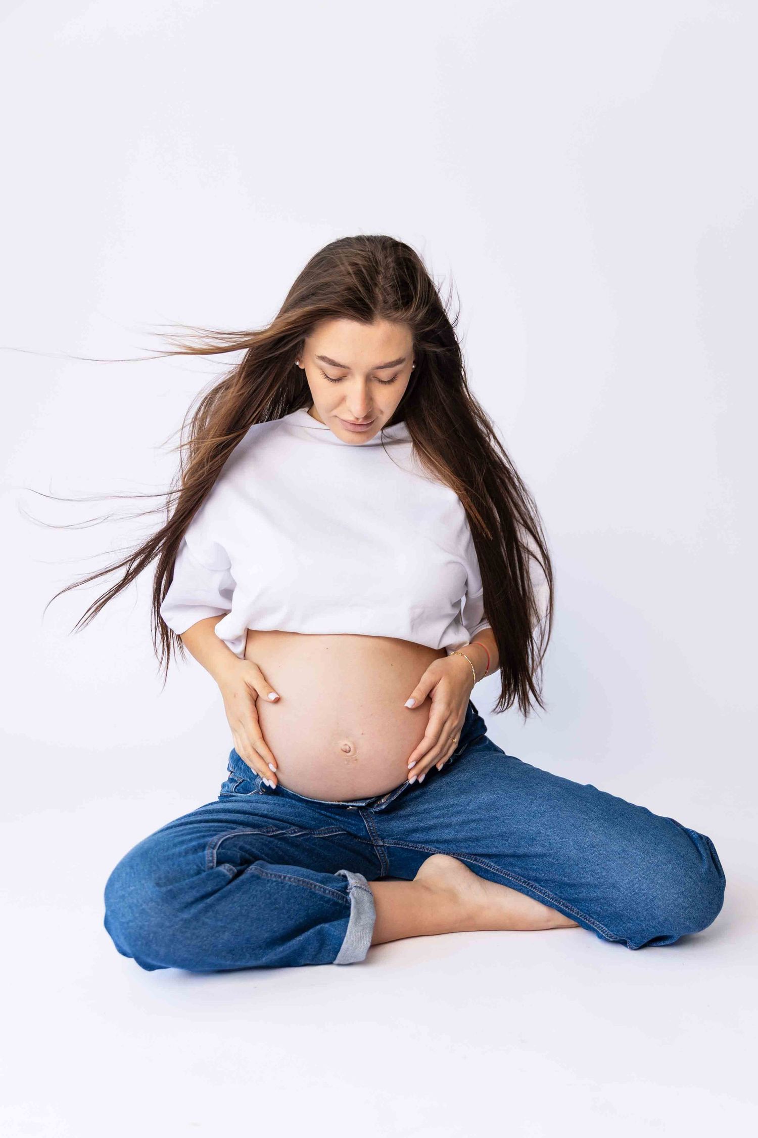 Pregnant person in white shirt and jeans sits cross-legged while gently touching belly as hair flows in breeze against white backdrop.