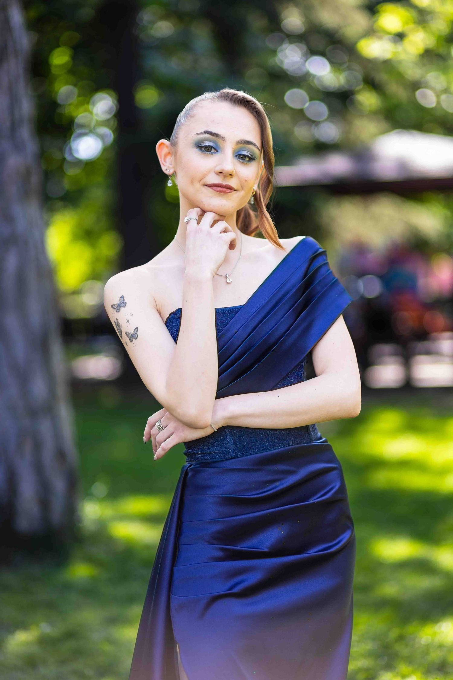A woman in a navy blue formal dress poses gracefully in a sunny outdoor park setting.