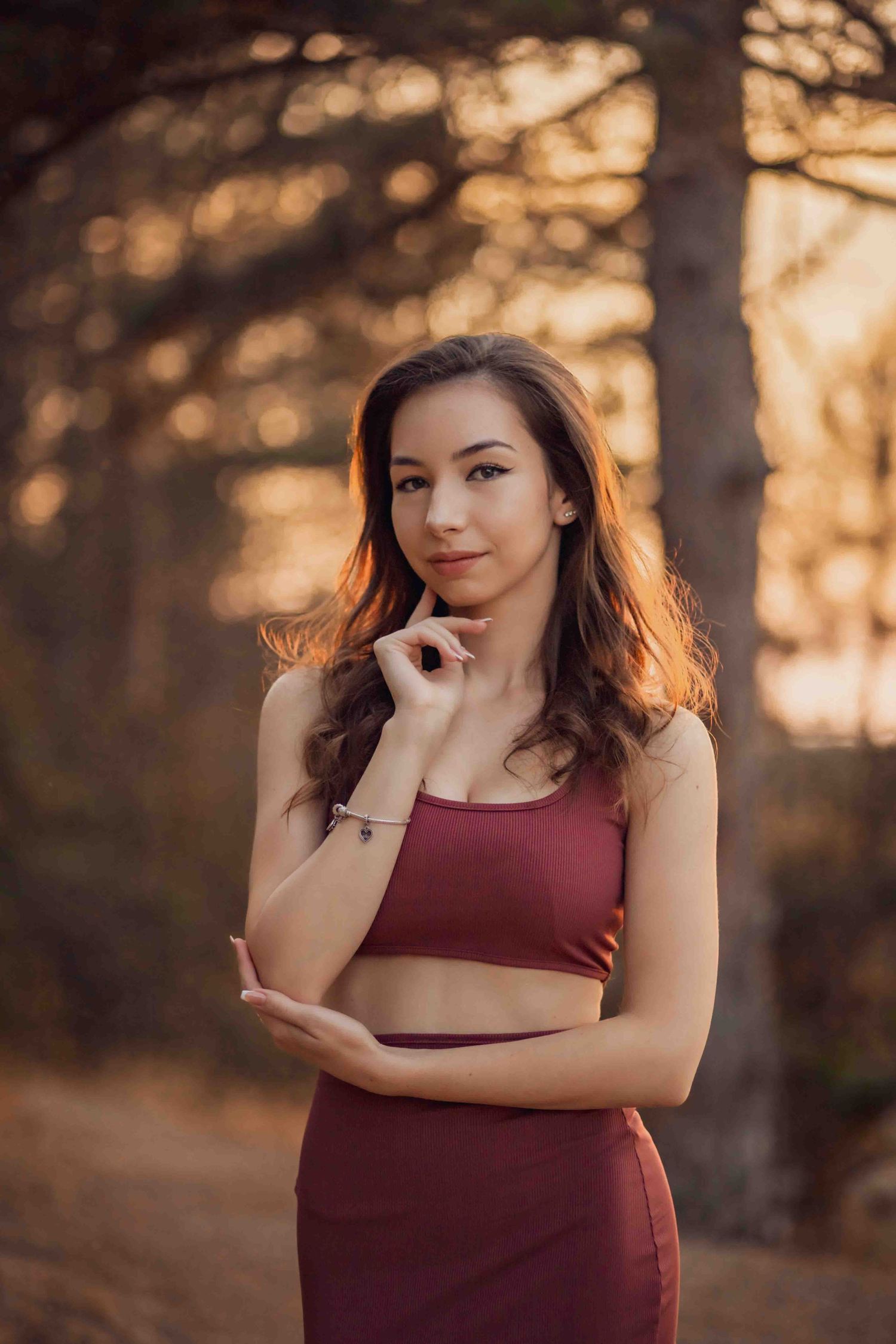 Athletic figure in burgundy workout set poses thoughtfully in golden sunset forest light.