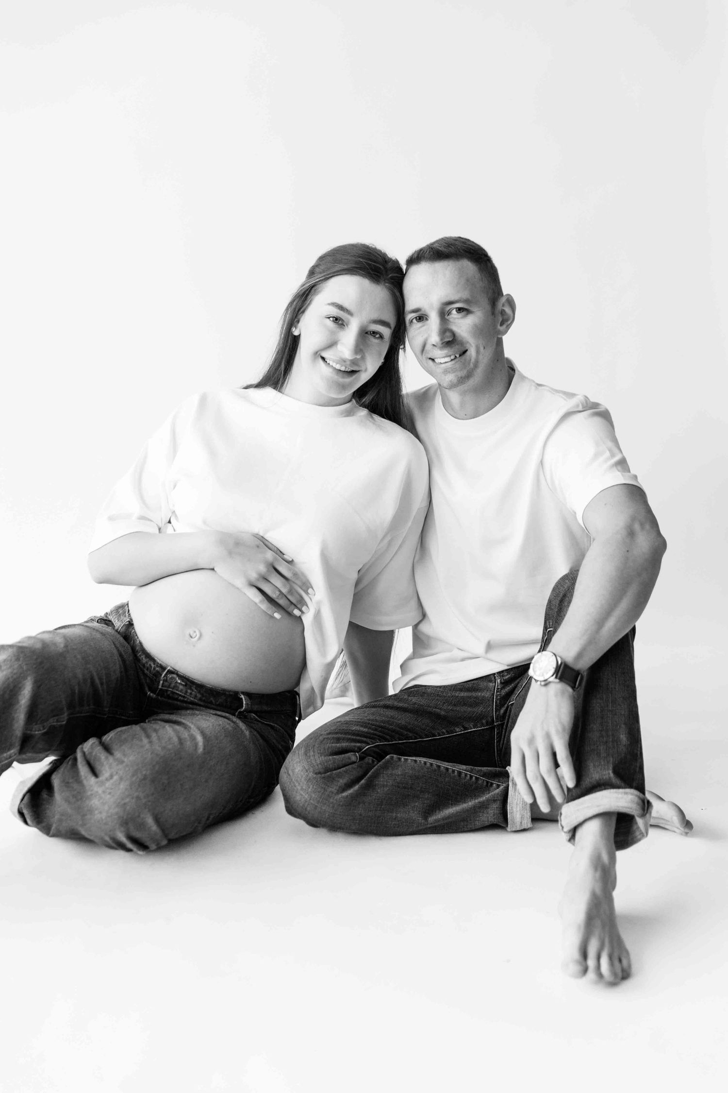 Black and white portrait of expecting couple in white shirts and jeans sitting together on seamless backdrop.